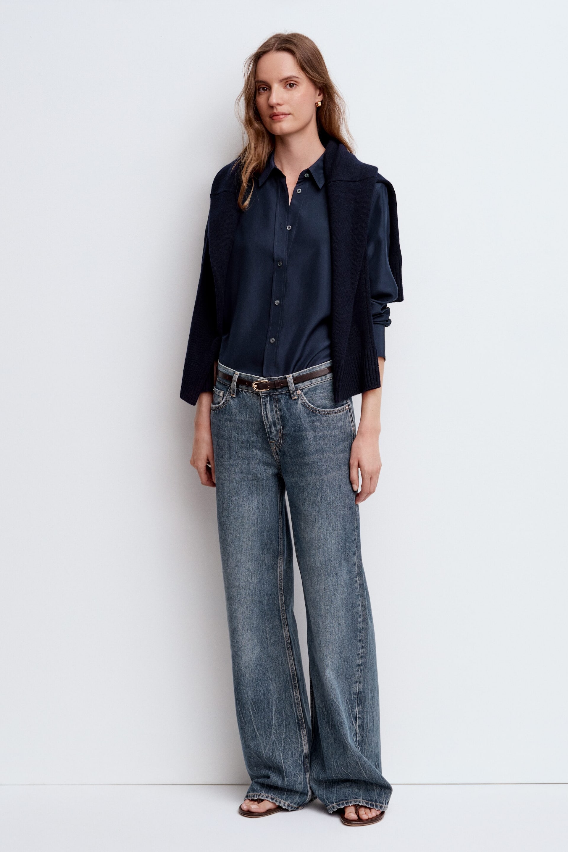 Visualizza immagine più grande: Una camicia blu navy a maniche lunghe con bottoni è indossata e infilata in jeans blu a gamba larga con lavaggio sbiadito e vita ben visibile sul davanti. Un maglione blu navy è adagiato sulle spalle, e una sottile cintura marrone stringe la vita.