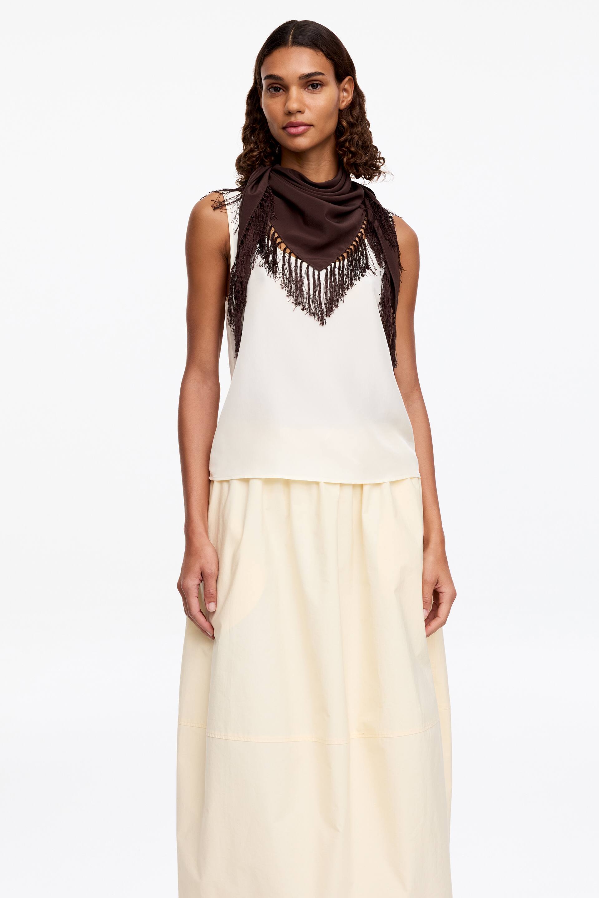 Преглед на по-голямо изображение: Копринен шал с ресни. Тъмнокафявият копринен шал, с декоративни ресни и малки мъниста, е носен от модела, преметнат около врата ѝ, като триъгълната му точка лежи върху гърдите ѝ над кремаво безръкавно горнище.