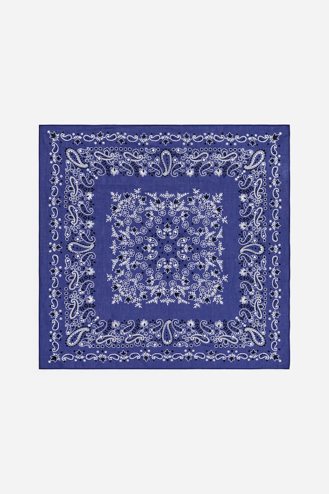 A blue bandana lies flat, displaying a white and black paisley pattern. The pattern features a central square of white floral and leaf motifs, surrounded by a border of white and black paisley teardrops and dots. An outer border of repeating smaller floral and paisley elements frames the design. - 1