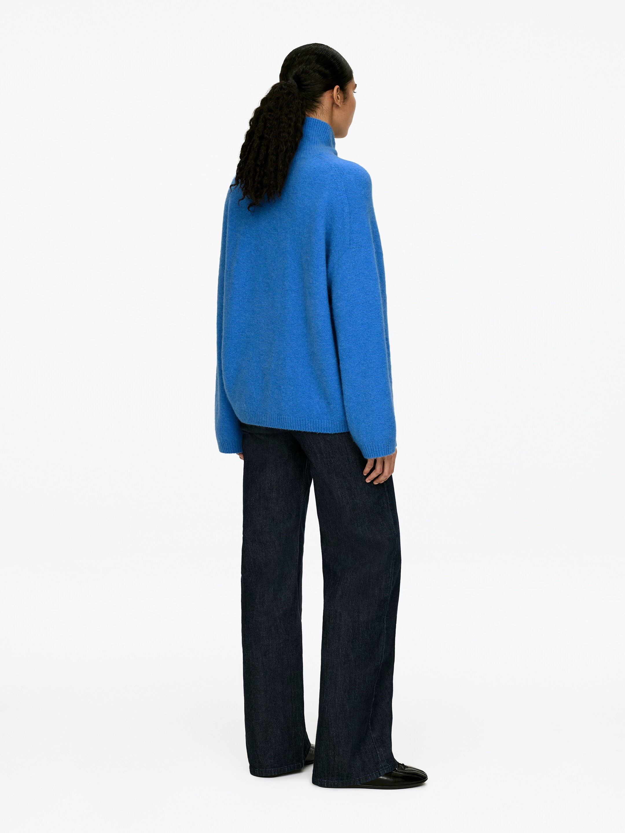 Vis større bilde: En blå, avslappet turtleneck-genser vises fra baksiden, med en høy hals og lange, løse ermer, og den bæres med mørke, vide bukser.