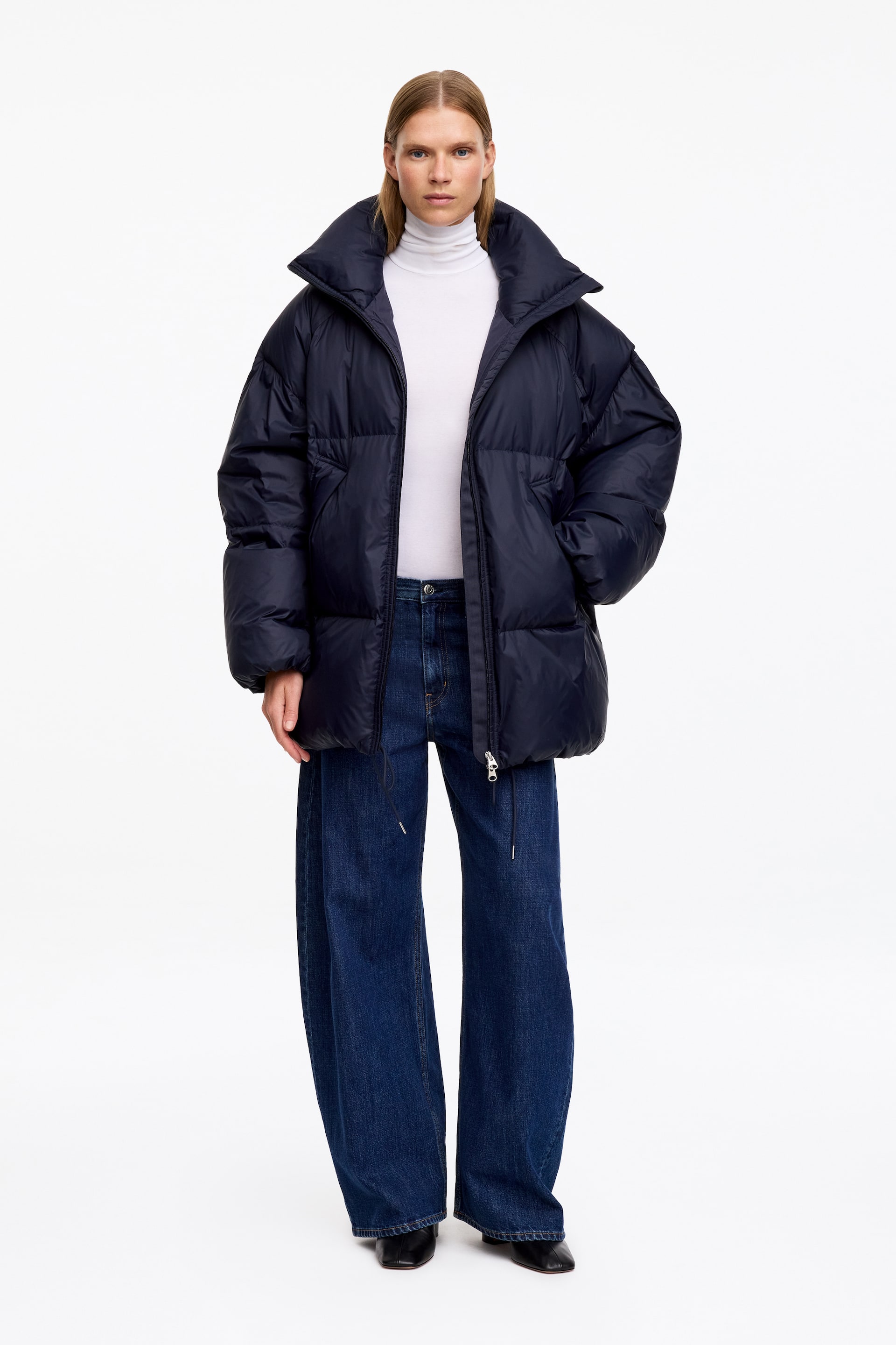 Visualizza l'immagine ingrandita: Una giacca puffer in piumino upcycled di colore blu scuro è indossata dalla modella, con collo alto e design trapuntato. Le mani della modella sono appoggiate nelle tasche laterali, mentre sotto la giacca parzialmente aperta si intravede un dolcevita bianco.