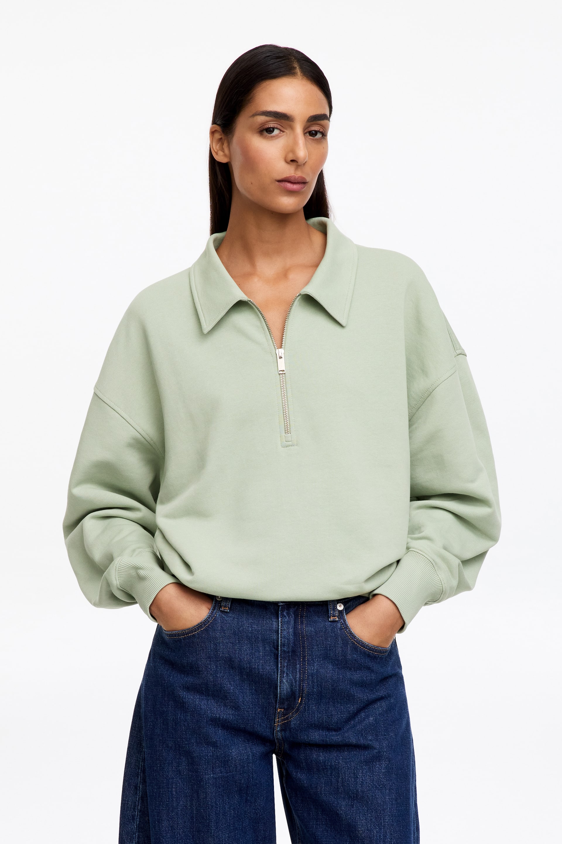 Visualizza immagine più grande: La modella indossa una felpa polo verde chiaro dalla silhouette rilassata. Presenta un colletto tipico, spalle scese, polsini a coste e una chiusura half-zip in finitura argentata con tiretto, parzialmente aperta. La modella indossa jeans blu scuro.