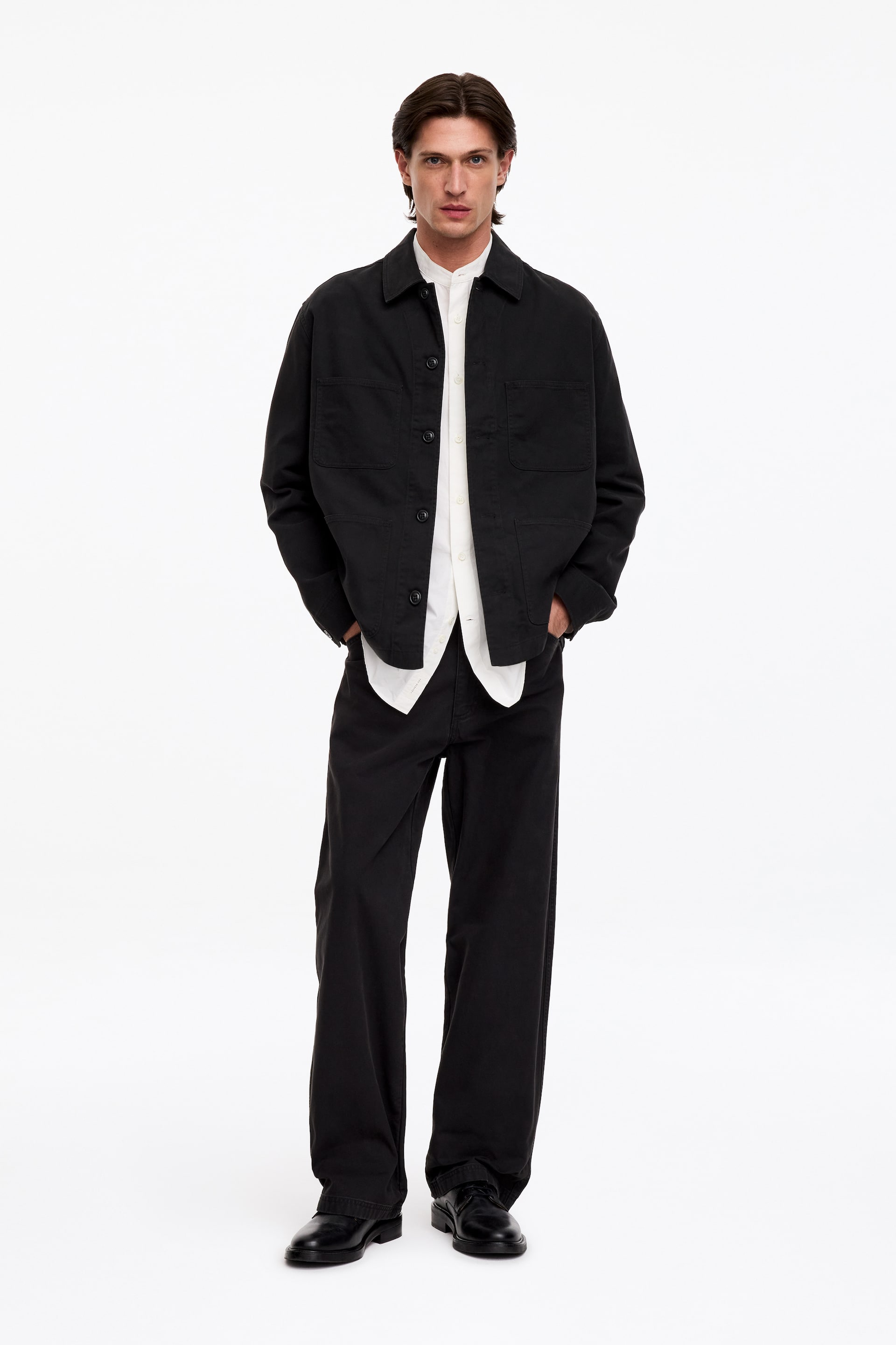 Größeres Bild ansehen: Ein schwarzes Utility-Overshirt wird vom Model getragen und über einem weißen Hemd mit Stehkragen kombiniert. Das Overshirt verfügt über einen Kentkragen, eine Knopfleiste vorn und zwei aufgesetzte Brusttaschen und wird mit einer schwarzen Wide-Leg-Hose sowie schwarzen Schnürschuhen gestylt, während das Model mit den Händen in den Hosentaschen steht.