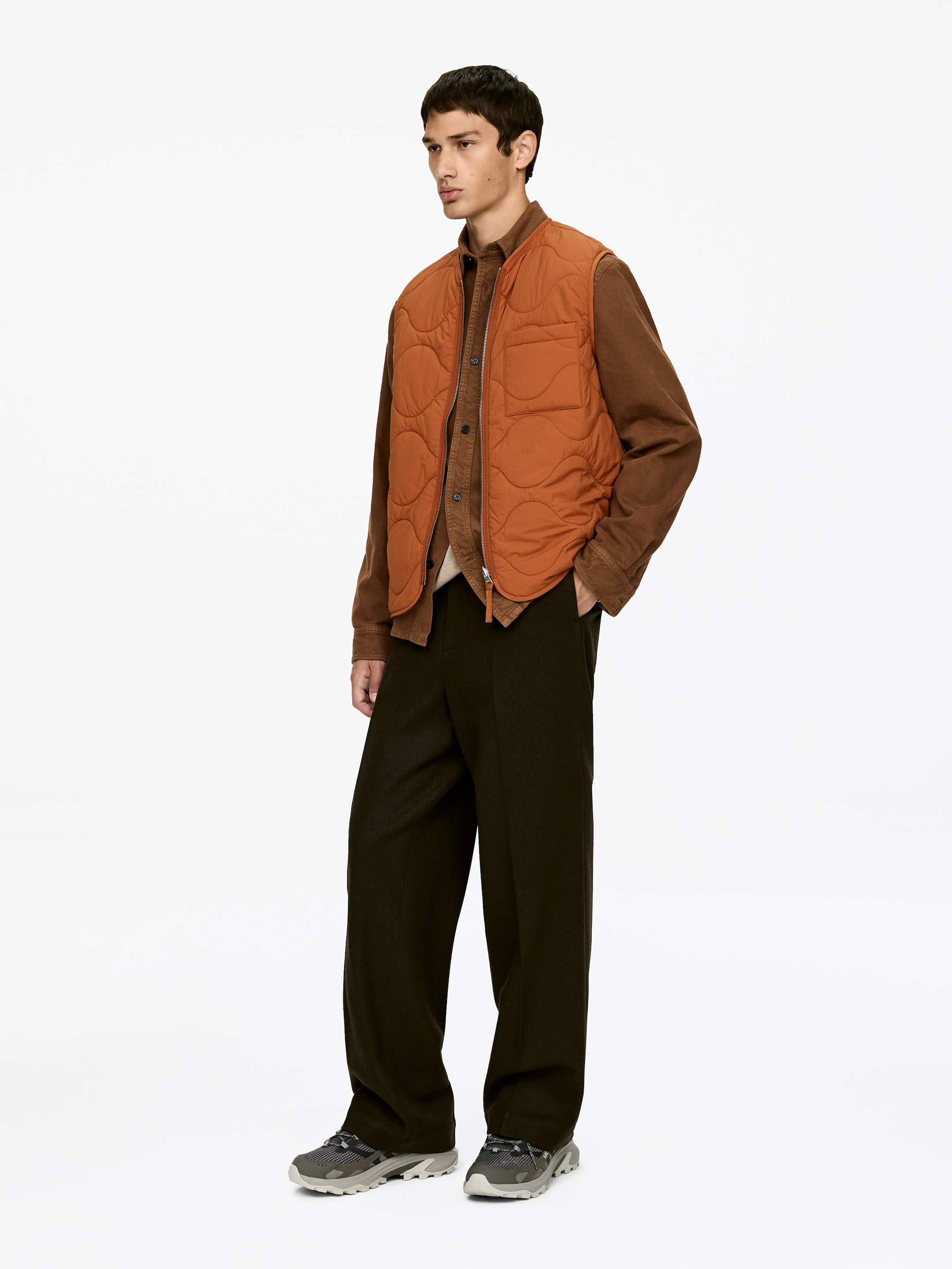Agrandir l'image: Un gilet matelassé orange, avec un motif de couture ondulé et une poche plaquée sur la poitrine, est superposé à une chemise marron à manches longues, assorti à un pantalon large marron foncé et des baskets épaisses grises et blanches. La tenue est présentée sous un angle légèrement orienté vers la droite.