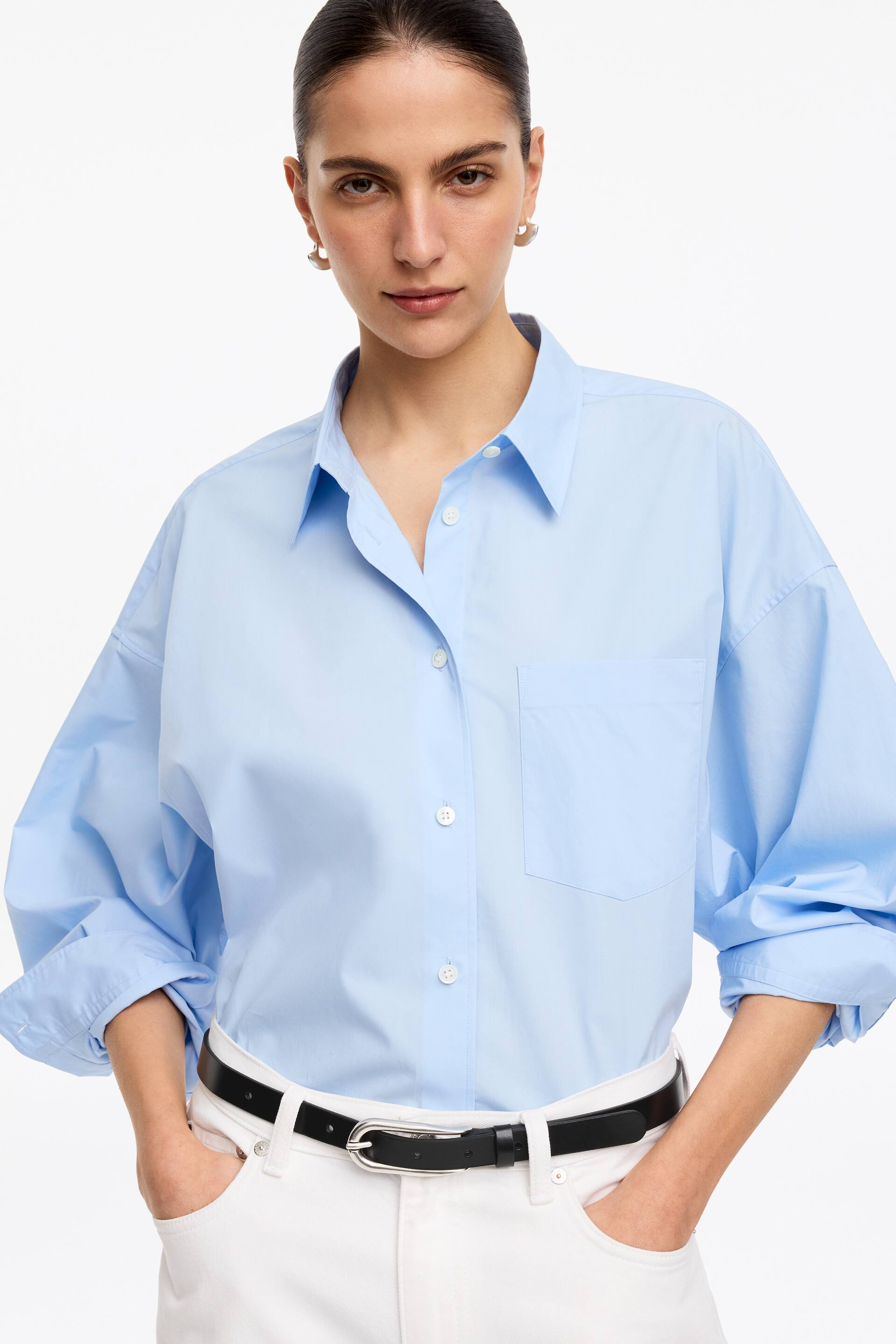 Agrandir l'image: Une chemise oversize bleu clair est présentée de face, avec un col pointu, des boutons blancs sur le devant et une poche plaquée unique sur la poitrine gauche. Ses manches longues et volumineuses sont retroussées jusqu'aux avant-bras, et elle est rentrée dans un pantalon blanc, révélant une ceinture noire.