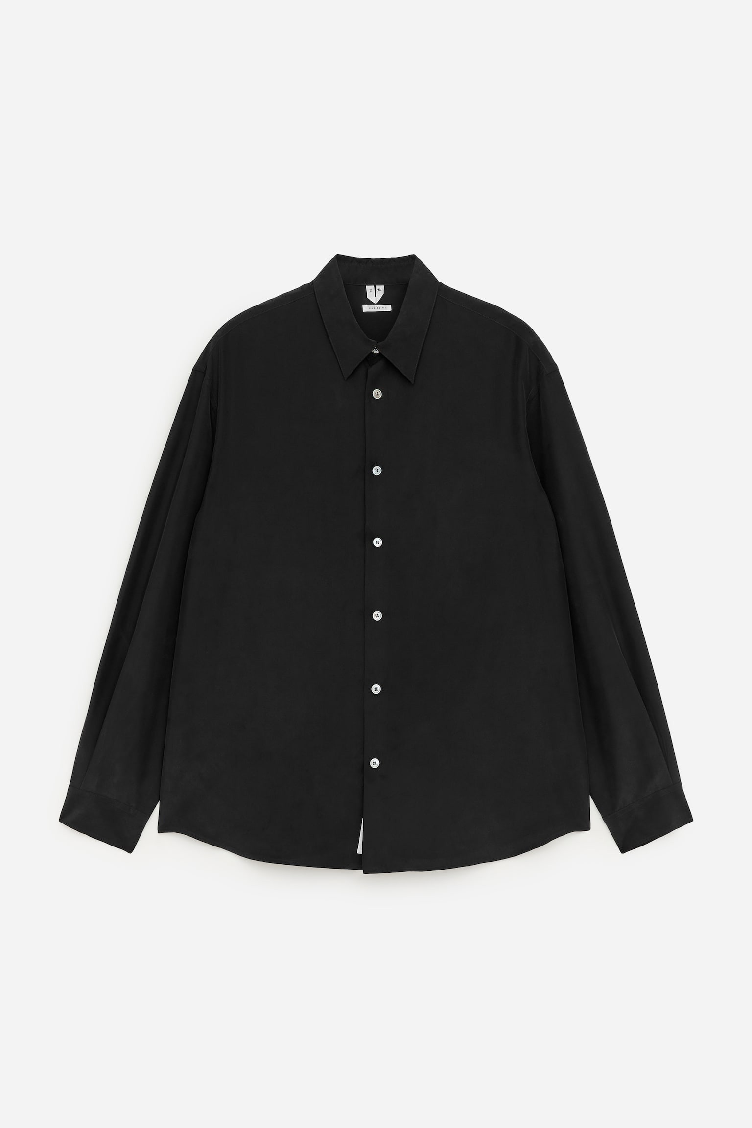 Una camisa de seda negra se presenta por delante, con un corte holgado, un cuello clásico y mangas largas con puños simples. Botones de madreperla blanca detallan la tapeta frontal, y un dobladillo ligeramente curvado completa el diseño.