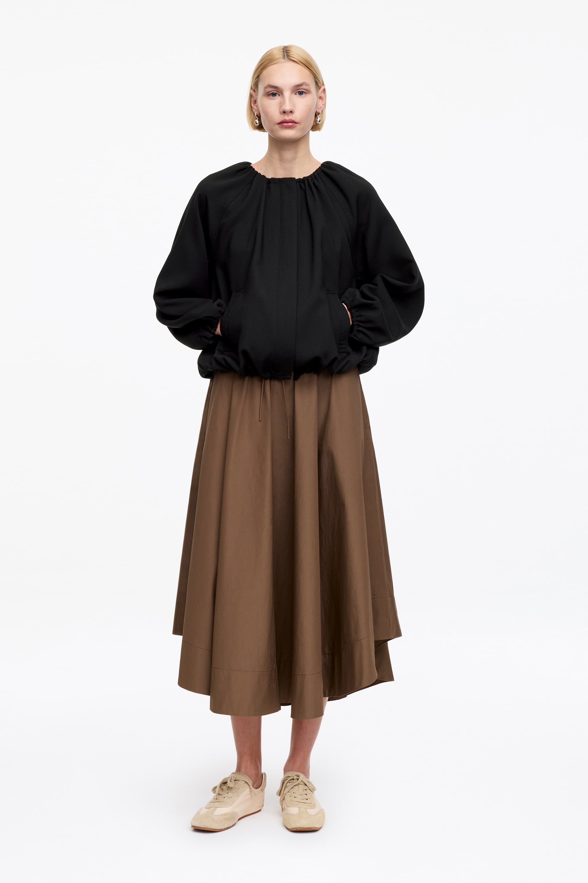 Näytä suurempi kuva: Musta blouson-paita esitetään edestäpäin, ja siinä on rypytetty pyöreä pääntie, runsaat pitkät hihat rypytetyillä kalvosimilla ja joustava helma. Tämä paita on yhdistetty ruskeaan A-linjaiseen midihameeseen, jossa on epäsymmetrinen helma ja näkyvä kiristysnauha vyötäröllä, kun taas vaaleanbeiget lenkkarit täydentävät kokonaisuuden.