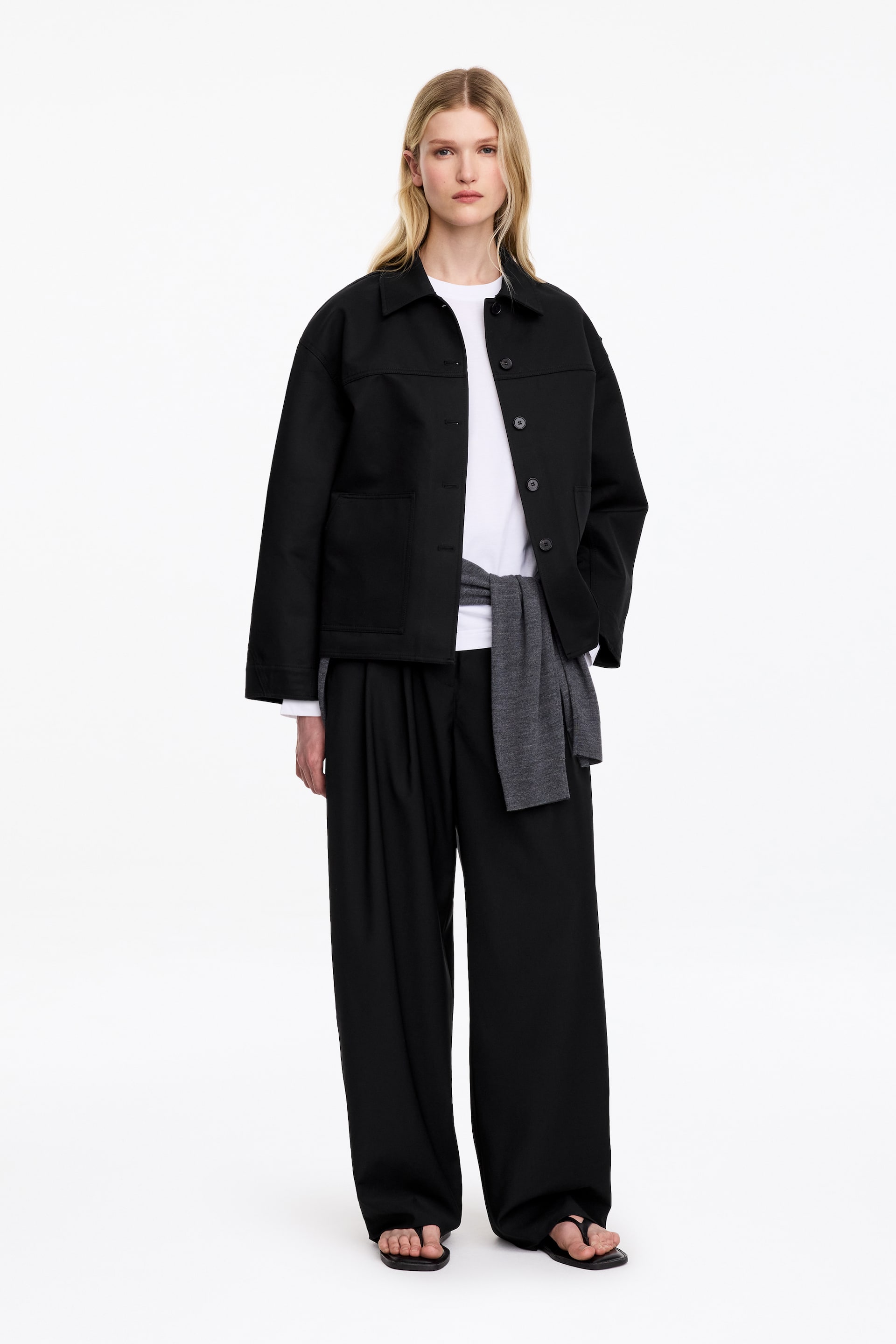 Visualizza immagine più grande: Una sovracamicia nera in cotone pesante, dalla silhouette strutturata ispirata al workwear, indossata aperta dalla modella. La sovracamicia è sovrapposta a una t-shirt bianca e abbinata a pantaloni neri a gamba ampia e a un maglione grigio annodato in vita.