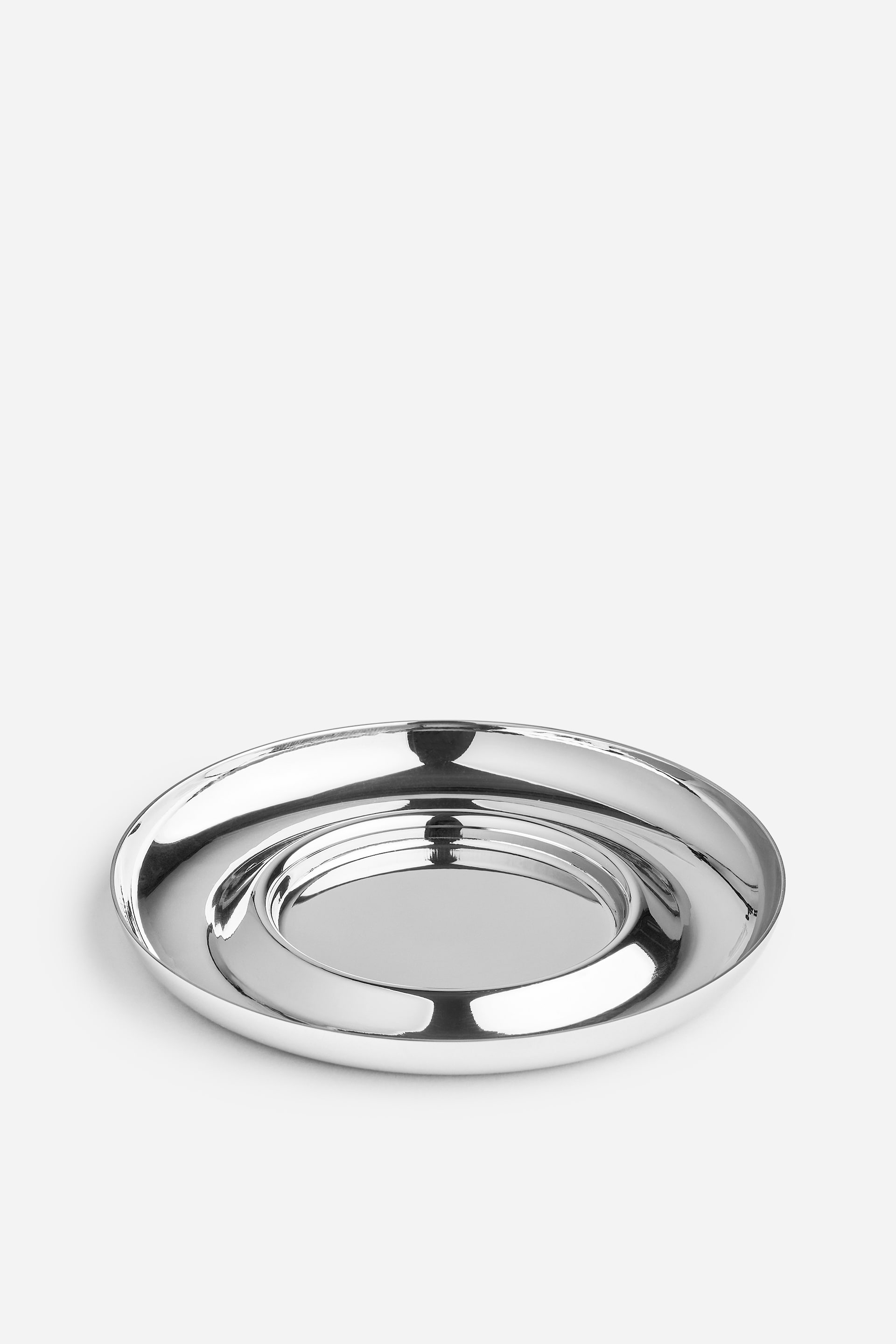 View larger image: A polished stainless steel saucer is resting at a slight front-facing angle, displaying its highly reflective metallic surface. The design features a shallow, raised outer rim and a circular recessed indentation in the centre.