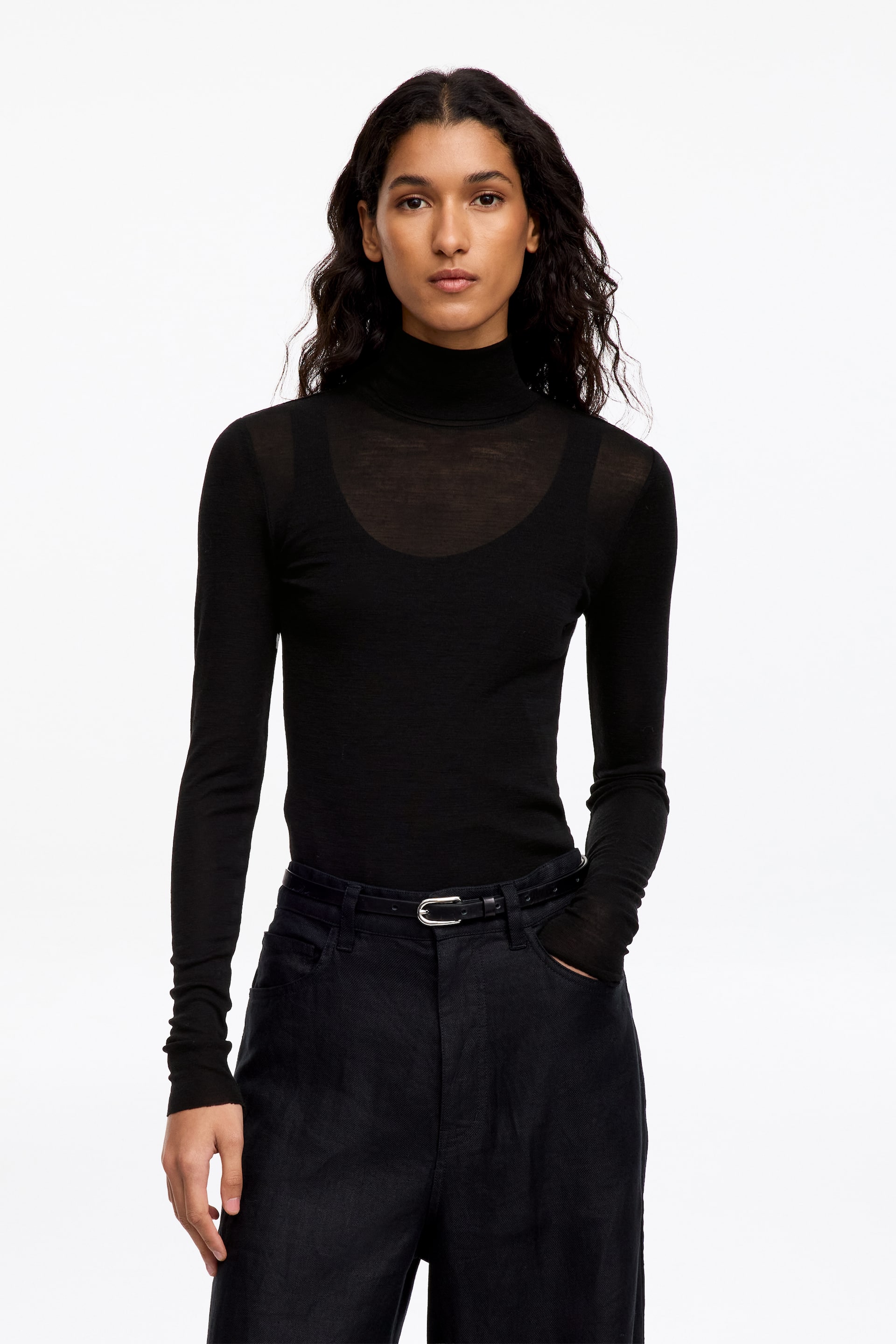 Se større billede: En sort, halvtransparent rullekraveskjorte med lange ærmer bæres af modellen, lagt over en matchende sort tanktop med rund hals. Den lette top er stukket ned i mørke bukser, som holdes på plads med et tyndt sort bælte.