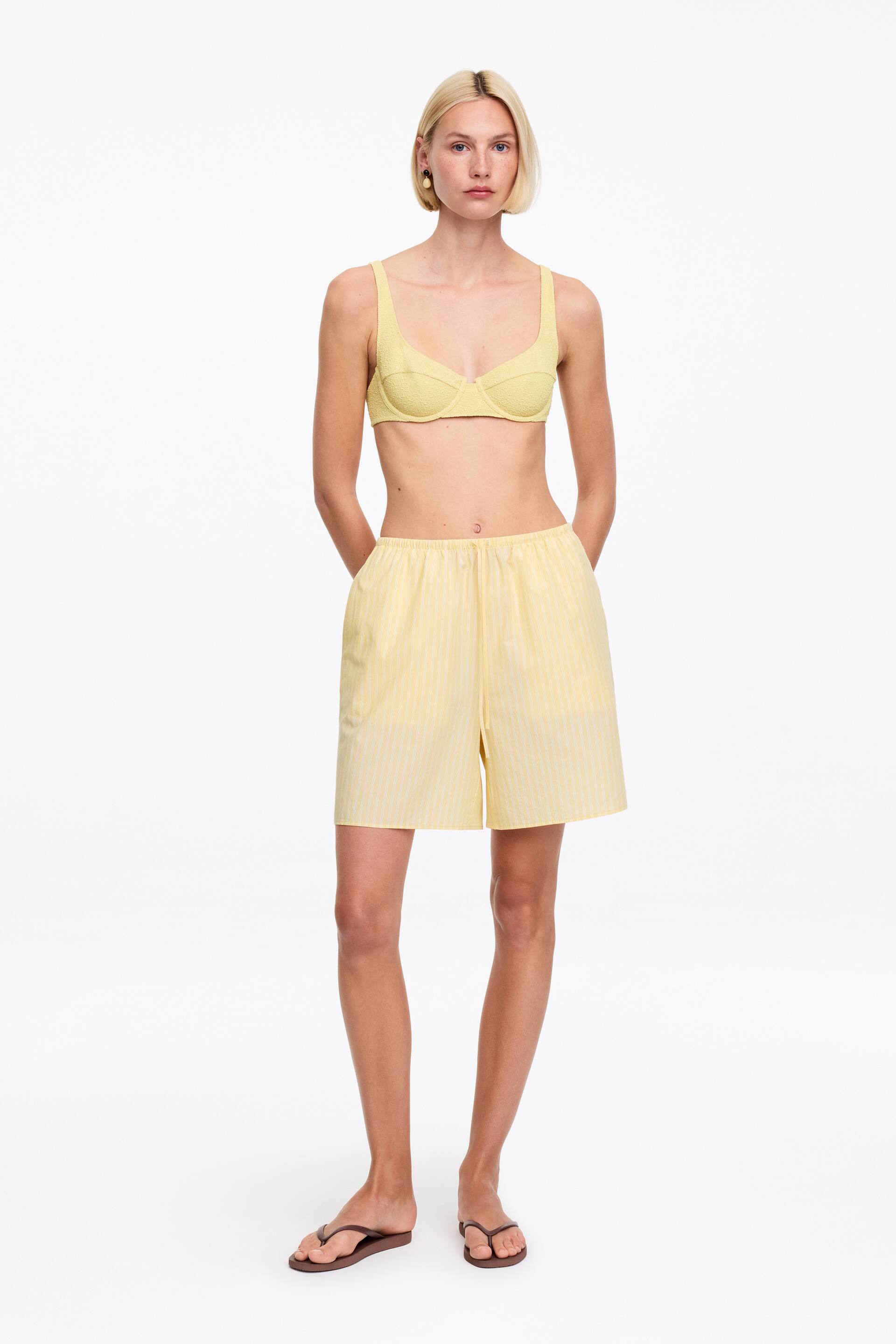 Agrandir l'image: Un haut de soutien-gorge texturé jaune avec des bonnets à armatures et des bretelles est présenté de face, assorti à un short jaune avec des rayures verticales subtiles et des poches latérales.