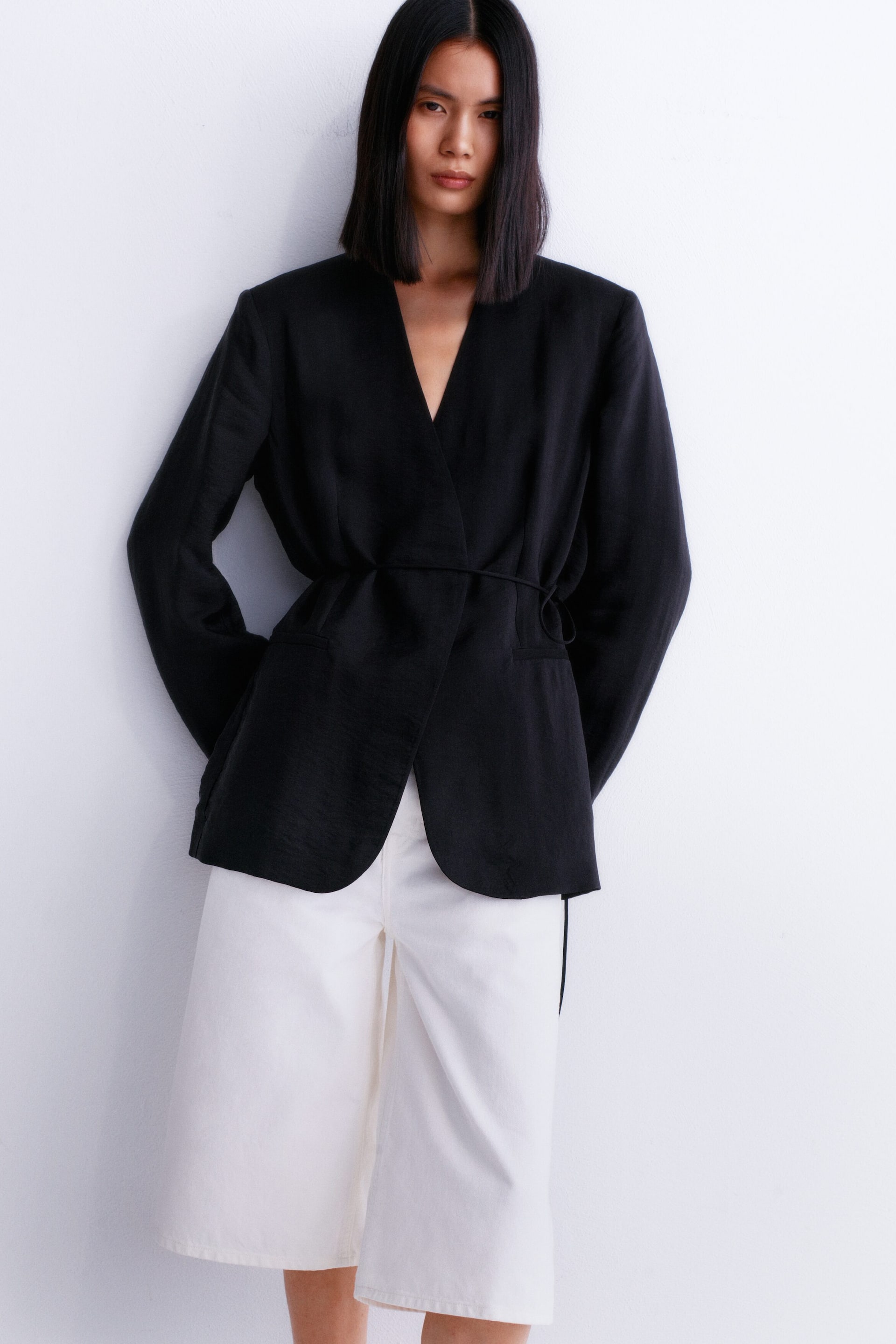 Ver imagen más grande: Una chaqueta negra cruzada, confeccionada en un tejido texturizado y con un profundo escote en V, mangas largas y un cinturón de lazo que ajusta la cintura, se lleva sobre unos pantalones cortos blancos de pierna ancha hasta la rodilla.