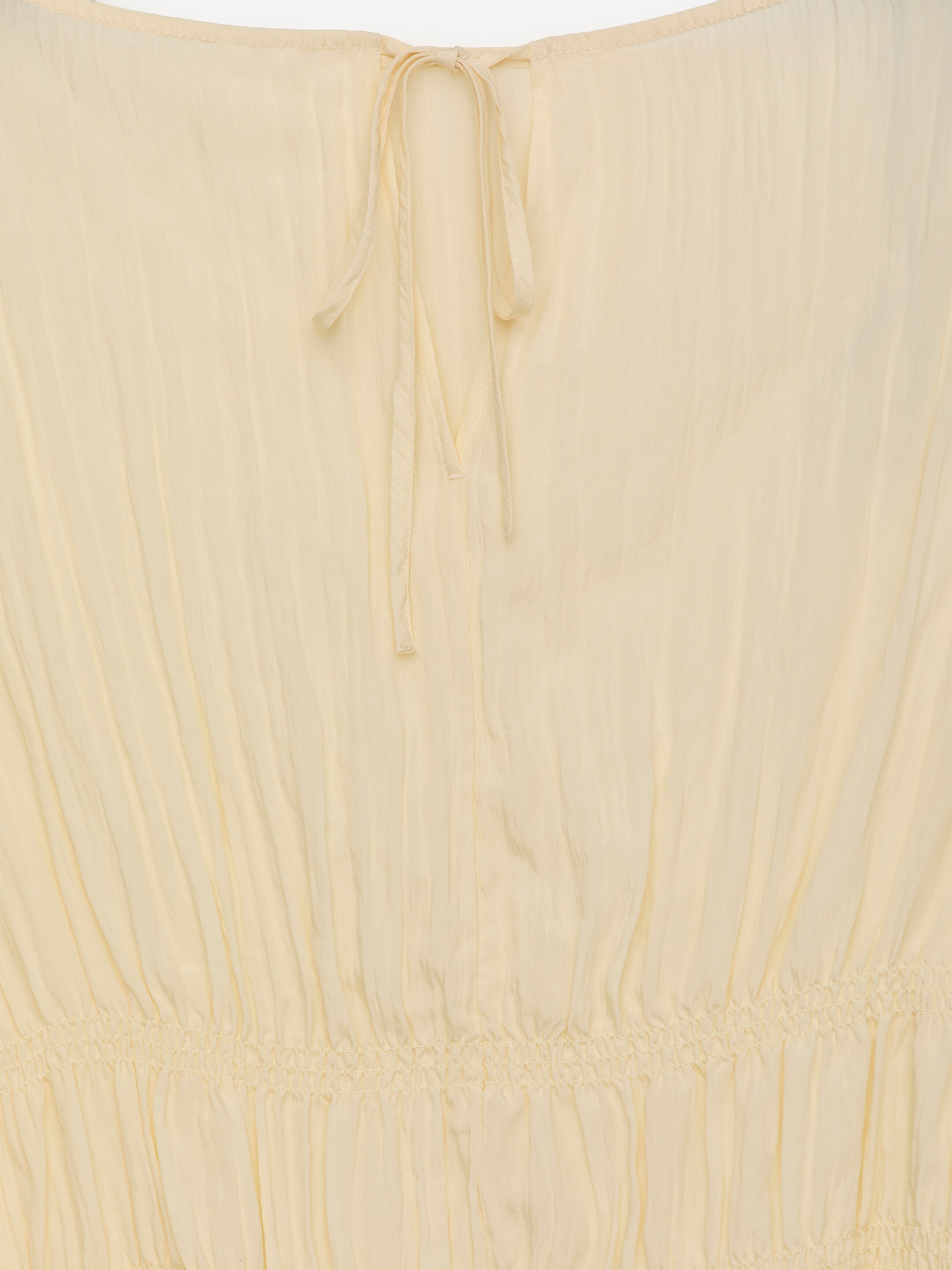 View larger image: A light yellow garment is presenting its back, featuring a knotted tie closure at the neckline above a V-shaped opening. Below, a horizontal gathered seam is cinching the fabric, creating vertical pleats throughout the garment.