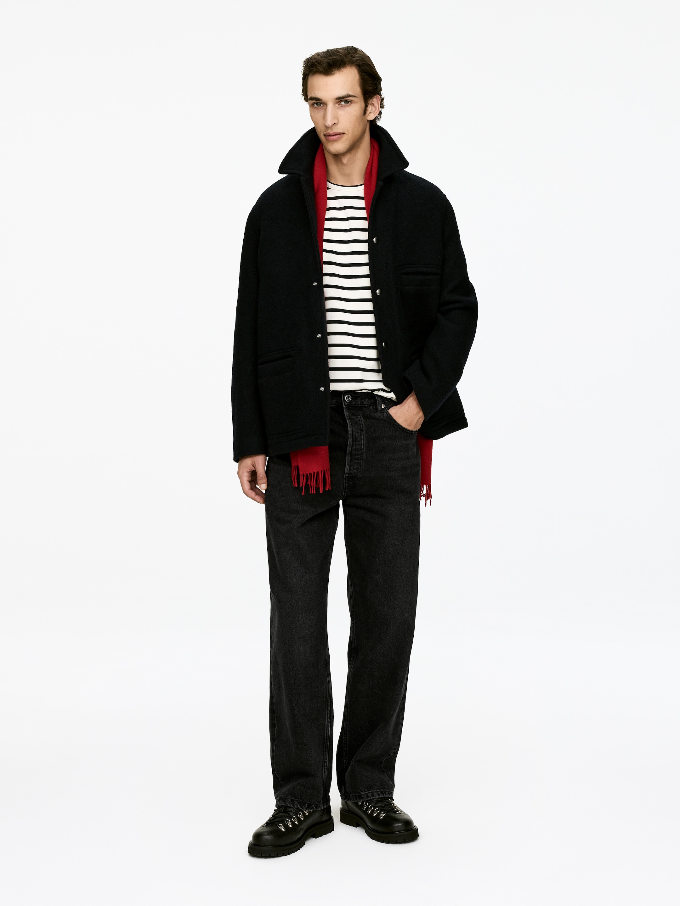 Grösseres Bild ansehen: Ein nach vorne gerichtetes Model trägt eine schwarze, strukturierte Jacke mit einem breiten Kragen und Druckknopfverschlüssen, geschichtet über einem weiß-schwarz gestreiften Langarmshirt und einem roten Fransenschal. Schwarze, locker sitzende gerade Jeans werden mit schwarzen Schnürstiefeln getragen, die mit metallenen D-Ring-Ösen und einer profilierten Sohle ausgestattet sind.