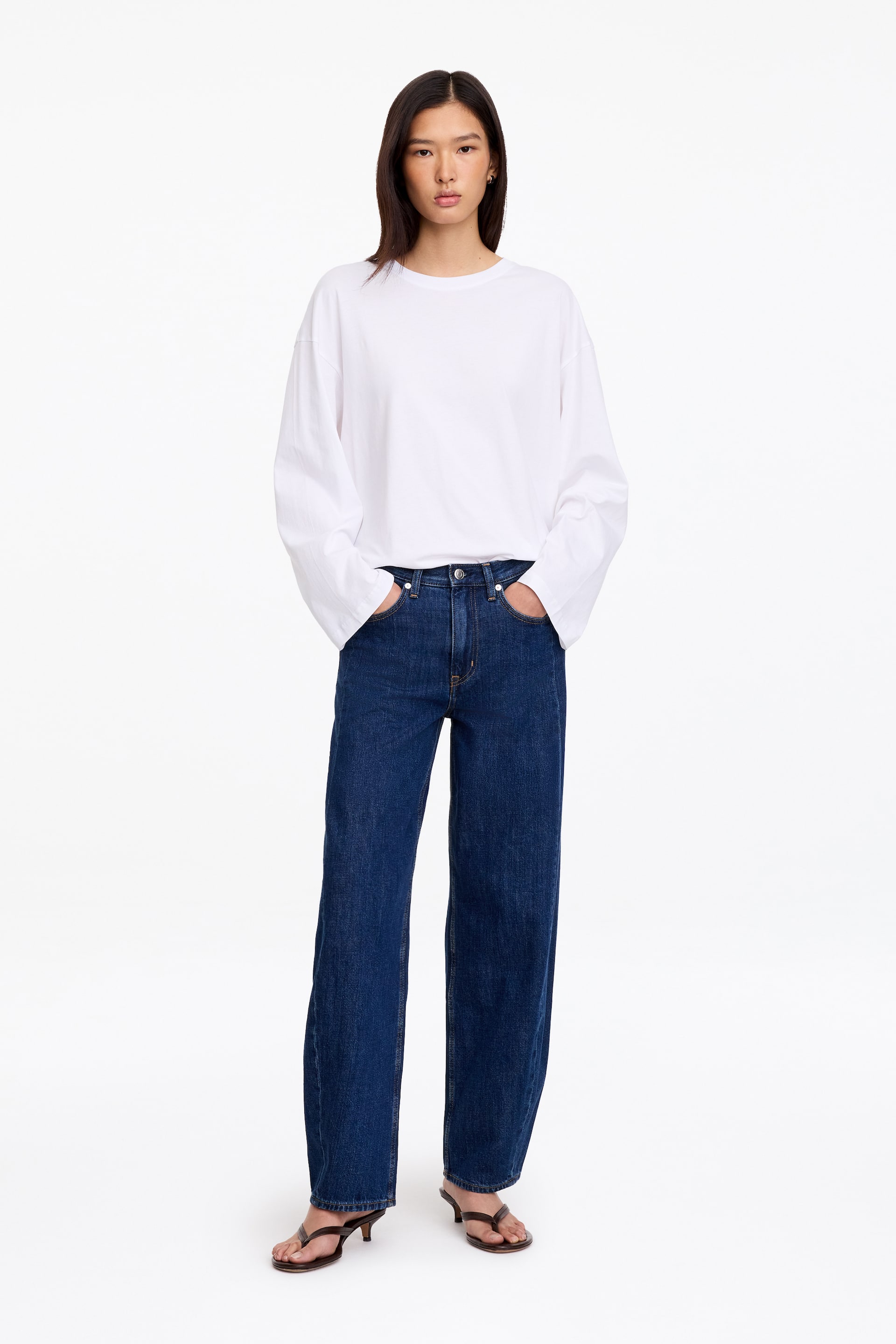 Grotere afbeelding bekijken: Donkerblauwe denim jeans met een relaxed fit en gevormde pijpen worden gedragen door het model, dat rechtop staat met haar handen in de voorzakken. De jeans zijn gestyled met een los wit T-shirt met lange mouwen en donkere sandalen met hak.