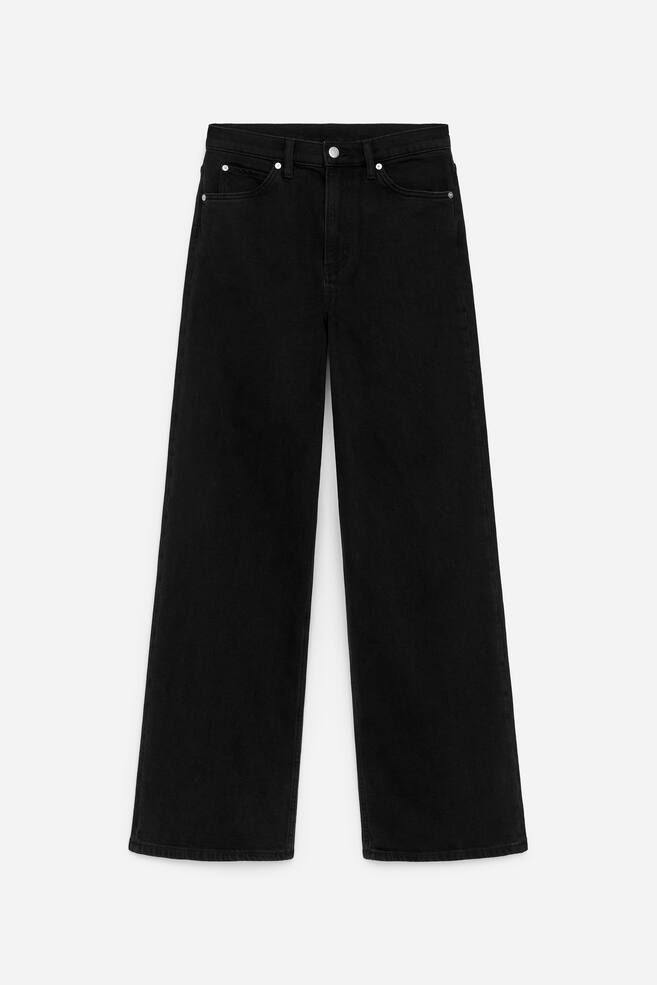 A pair of black denim jeans is being presented from the front, featuring a high-rise waist and a button closure for the zip fly. The jeans are designed with a slim fit through the hips, transitioning into wide, flared legs. - 1
