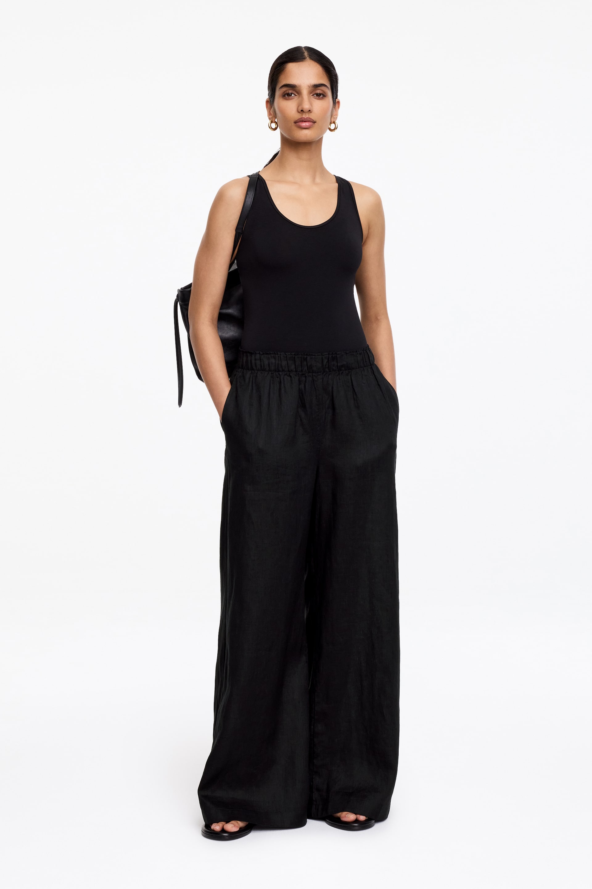 Se større billede: En sort ærmeløs top med scoop-hals sidder tæt og bæres stukket ned i sorte bukser med brede ben. Bukserne har en elastisk linning og viser et tekstureret stof, der flyder løst ned til gulvet.