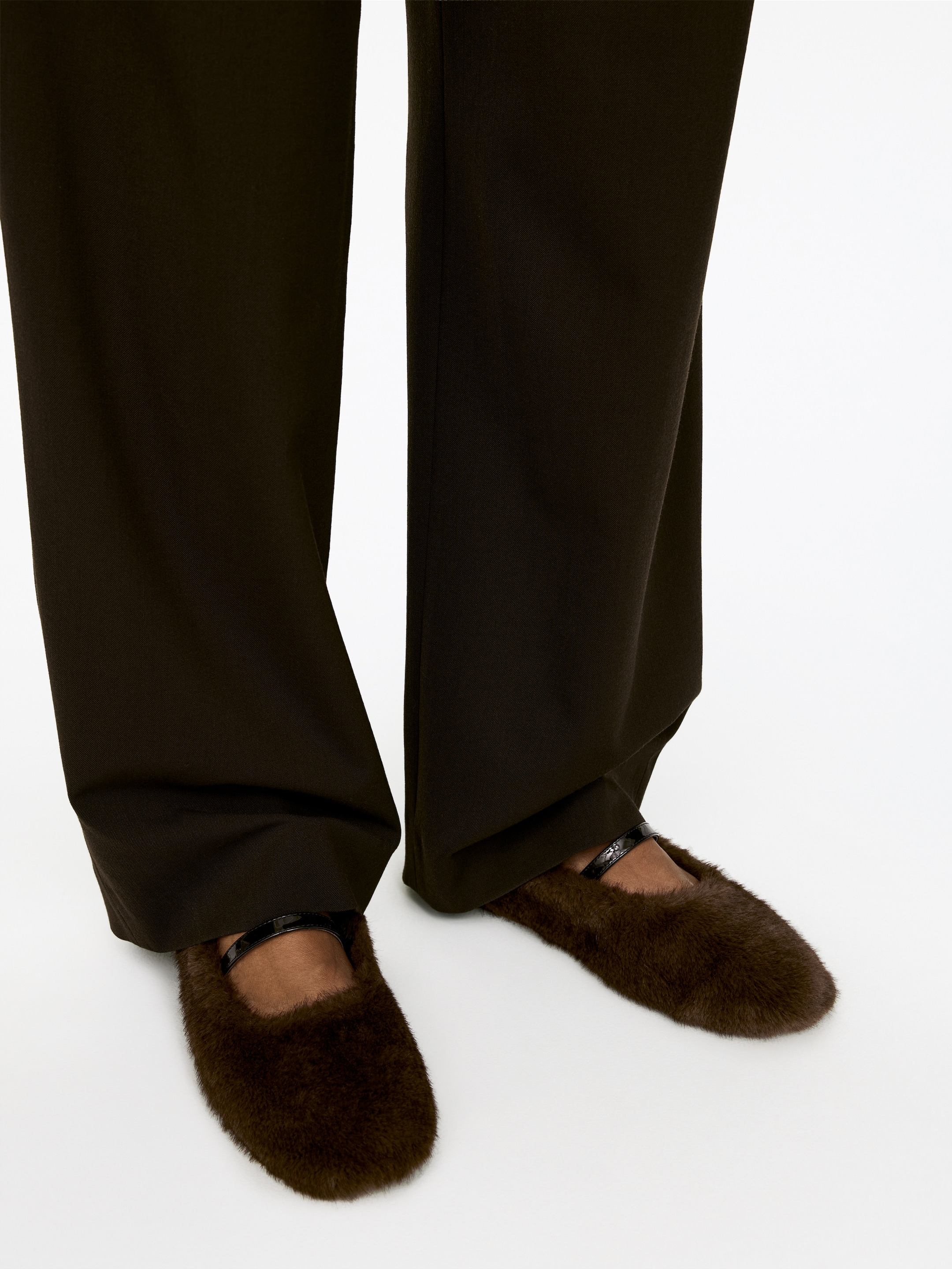 View larger image: Dark brown, plush Mary Jane shoes are being presented from a slightly elevated, front-facing angle. A black, textured strap is securing the shoes across the instep, while dark brown trousers are falling over their top edges.