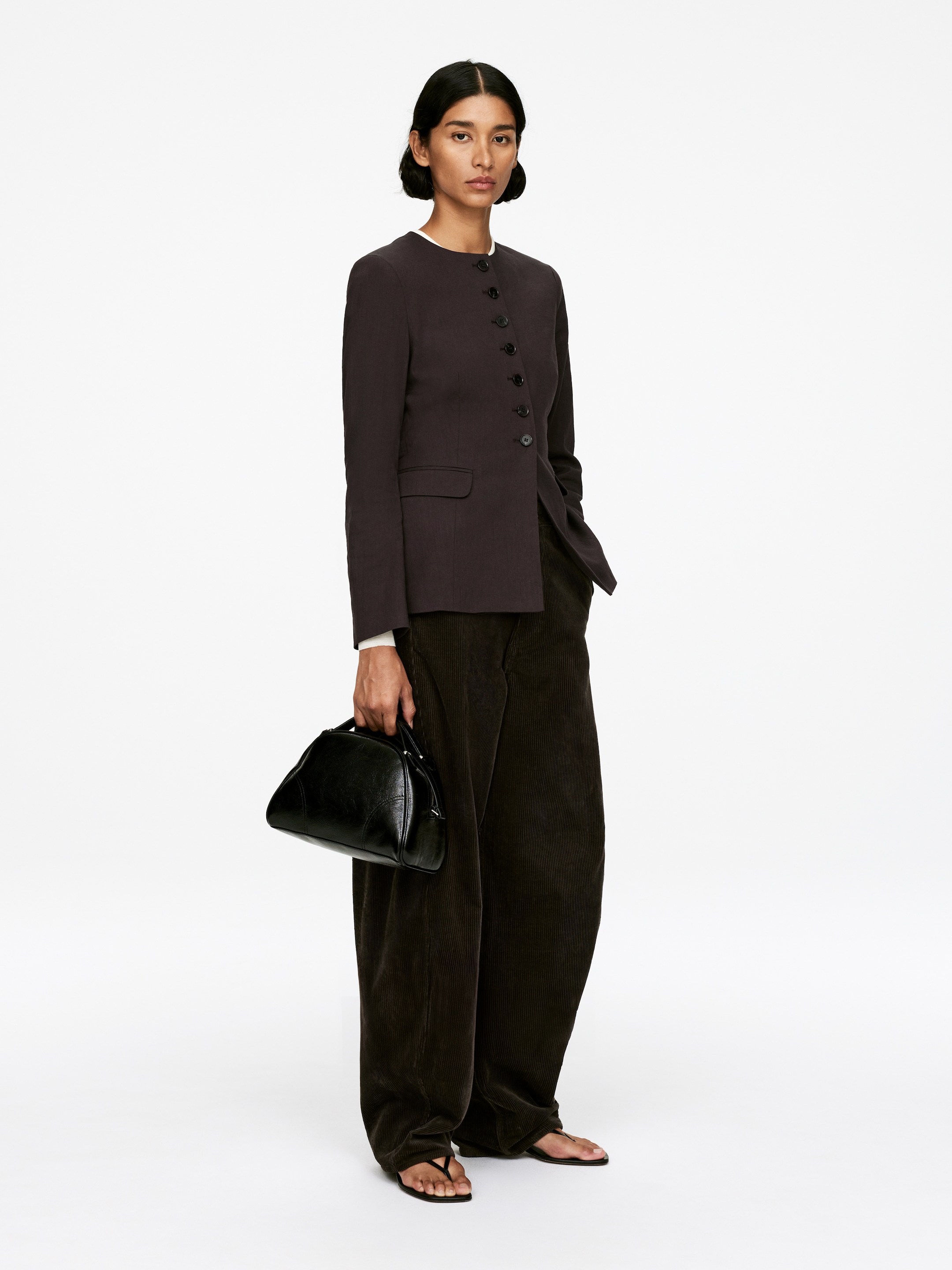 View larger image: A dark collarless blazer is being worn by the model, featuring an off-center button closure, subtle dart detailing, and a flap pocket. The blazer is cut for a slim fit and is being layered over a white top.