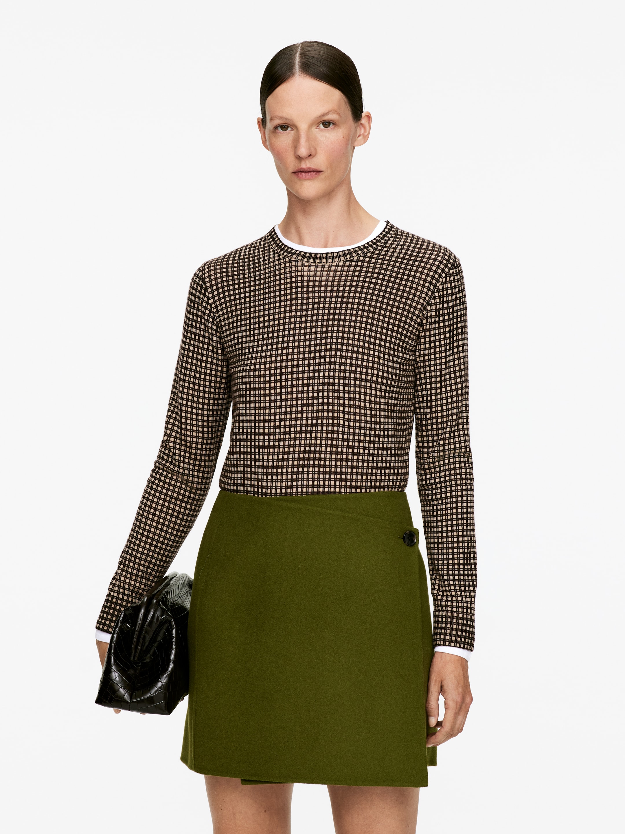 Se større billede: En slim-fit, langærmet top med et brunt, sort og lyst mikro-ternet mønster bæres over en hvid crew-neck top, synlig ved halsudskæringen og manchetterne. Den er parret med en olivengrøn A-line wrap-nederdel med en mørk knaplukning på højre hofte.