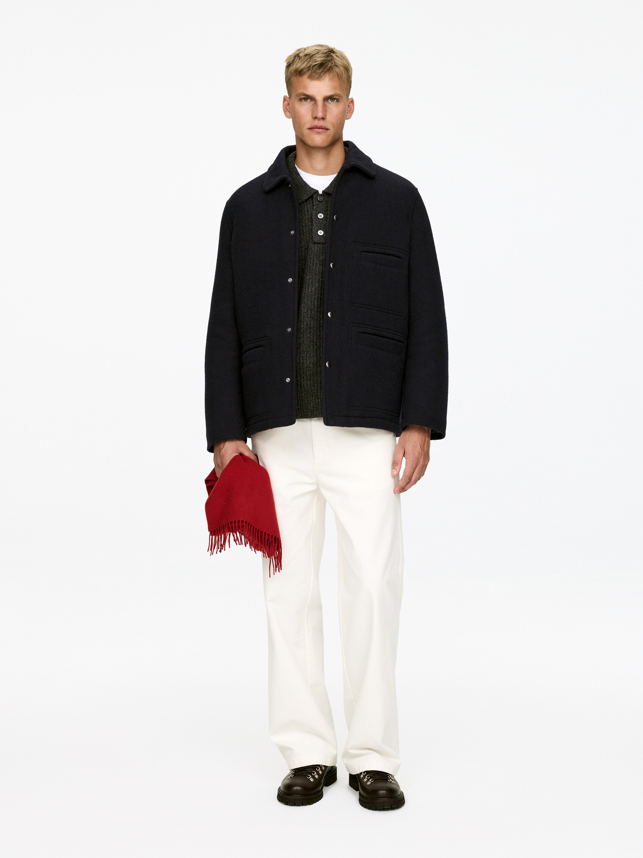 Преглед на по-голямо изображение: Тъмно синьо сако с текстура е представено с предна част, с яка на деколтето, закопчаване с копчета отпред и четири джоба на капаче. То е показано на пластове върху тъмно зелена плетена поло риза и бяла тениска, в комбинация с кремави панталони с широки крачоли, тъмни обувки с връзки и червен шал с ресни, който се държи.