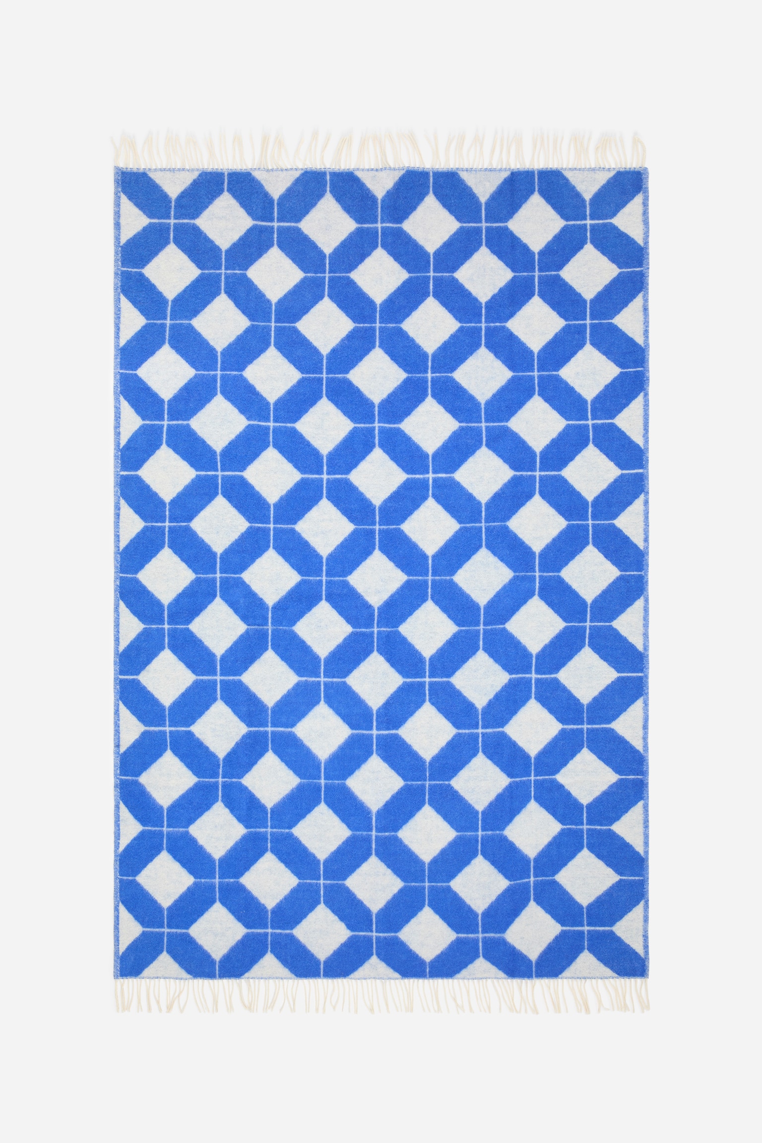 Eine rechteckige Wolldecke ist von vorne zu sehen und zeigt ein sich wiederholendes, blaues und cremefarbenes, jacquardgewebtes geometrisches Kachelmuster. An den kÃ¼rzeren oberen und unteren Kanten sind kurze, hellfarbene Fransen angebracht. - 1