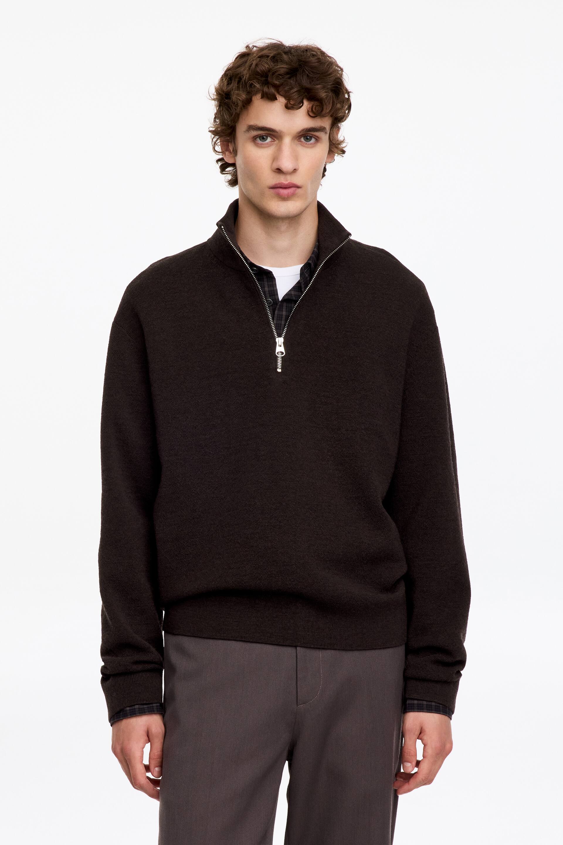 View larger image: A dark brown half-zip knit jumper is being worn by the model, layered over a dark plaid shirt and a white T-shirt. The jumper features a high collar with a silver zip, ribbed cuffs, and a ribbed hem, and is paired with grey trousers.