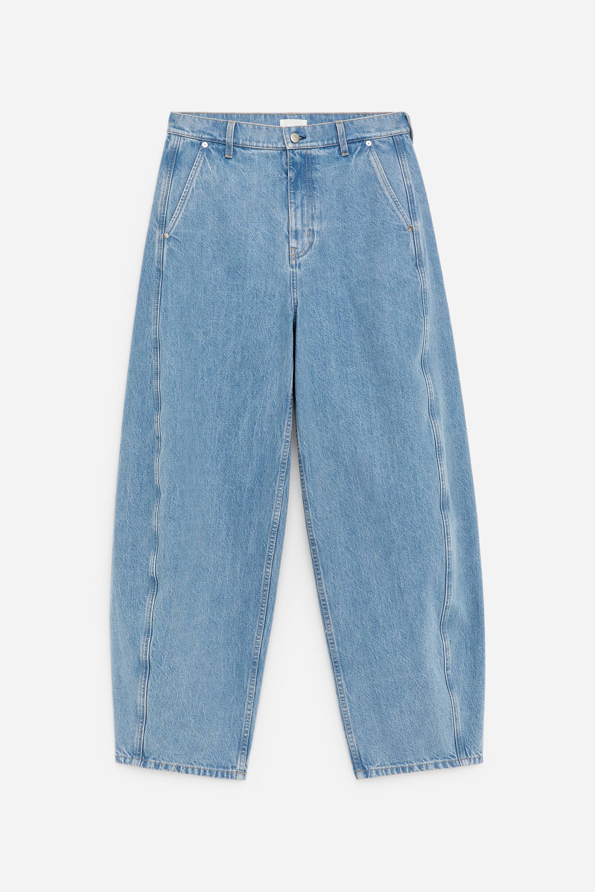 Se større billede: Tøndeformede denim bukser vises forfra, med en mellemhøj talje, skrå sidelommer og en lynlåslukning. En markant lodret søm løber ned langs forsiden af hvert ben, hvilket bidrager til tøndesilhuetten.