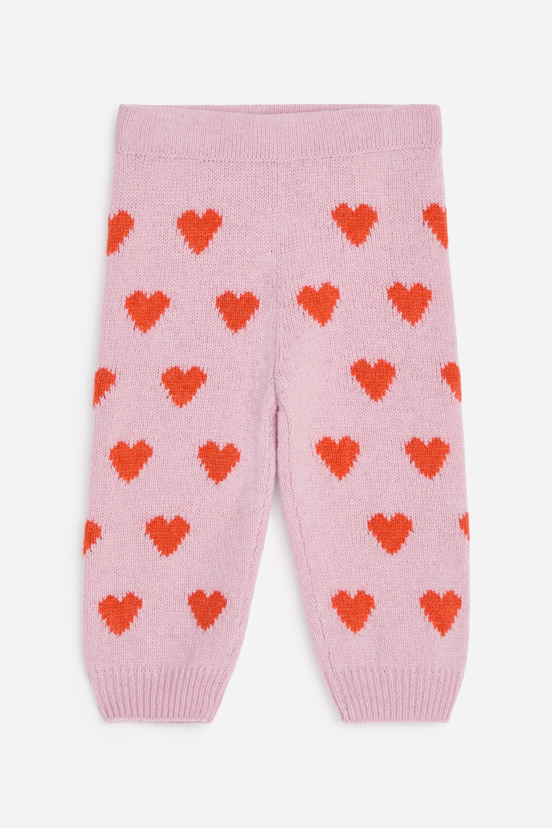 View larger image: These pink knitted trousers are shown from the front, featuring an all-over red heart pattern, a ribbed elasticated waist, and ribbed cuffs at the ankles.