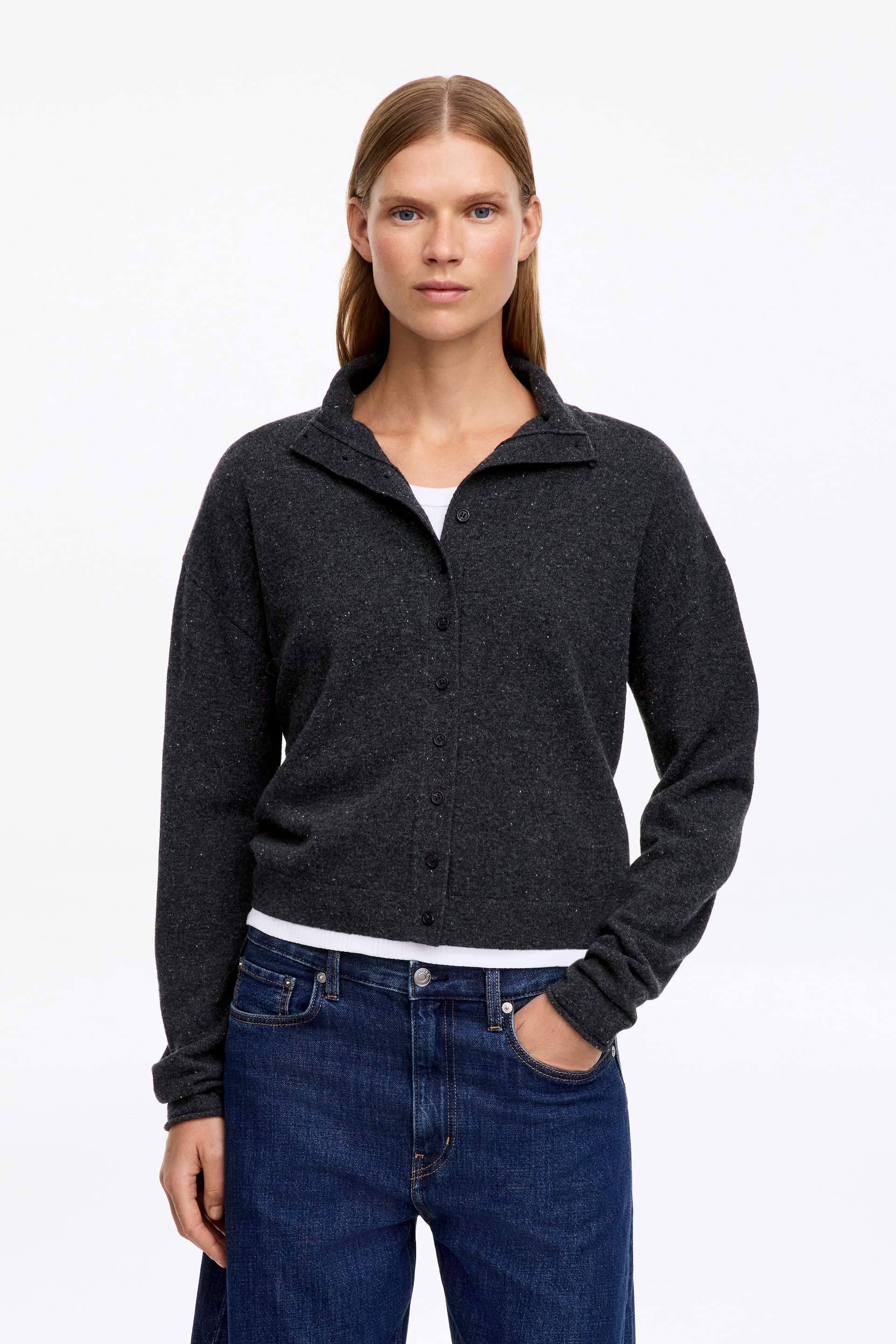 View larger image: A dark grey flecked knit cardigan featuring a button-through front and a softly structured chimney neckline is being worn by the model. The cardigan is styled layered over a white T-shirt and paired with blue jeans.