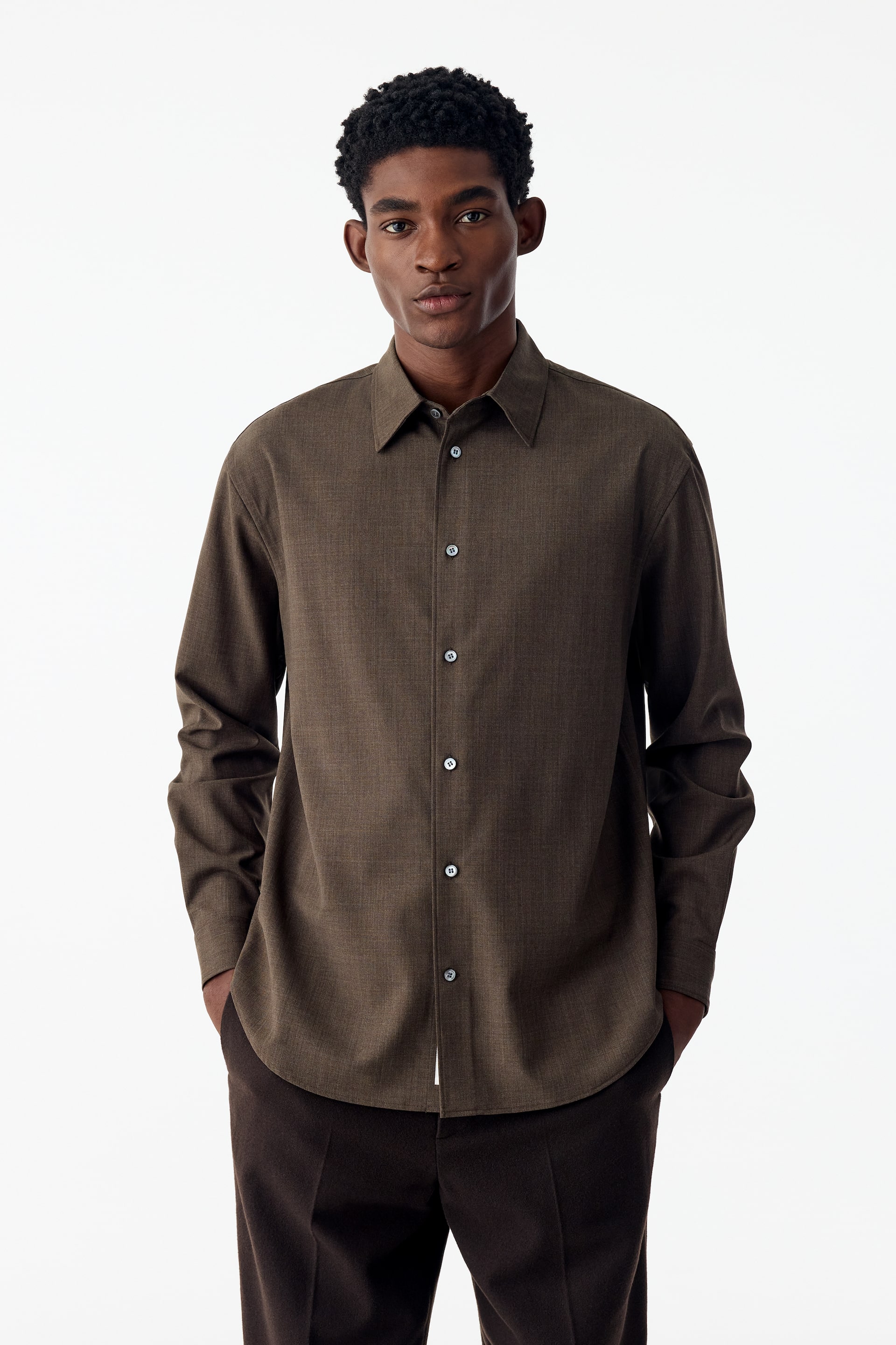 View larger image: The model is wearing a long-sleeved, relaxed-fit brown shirt with a point collar and button-up front. The shirt features a curved hem and is styled untucked over dark trousers.