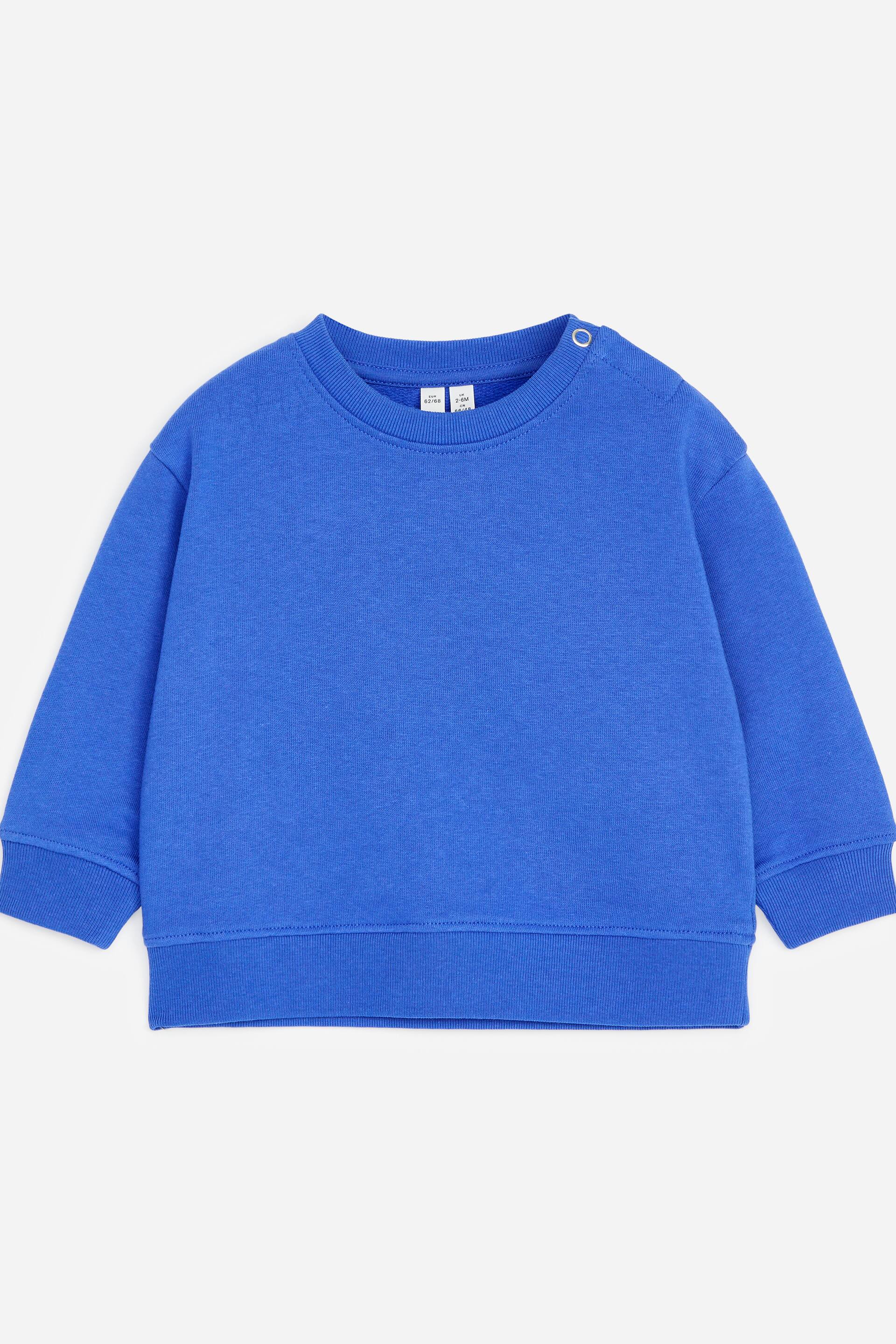 Grotere afbeelding bekijken: Een blauw sweatshirt met reguliere pasvorm toont zijn voorkant, met een ronde halslijn en een enkele drukknoopsluiting op de linkerschouder, en geribde boorden en zoom.