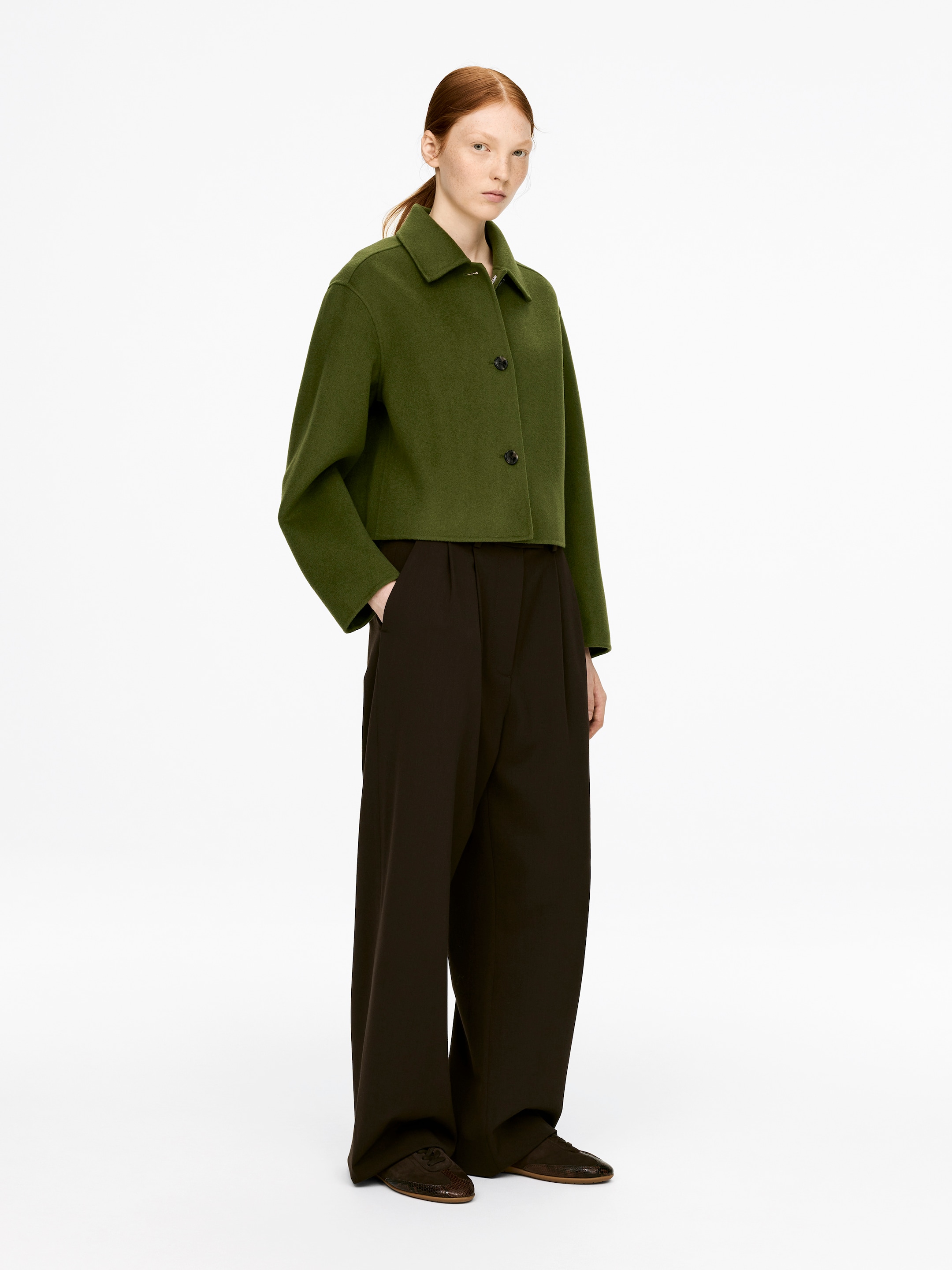 Ver imagen más grande: Una sobrecamisa verde oliva con cuello y tres botones está siendo llevada por el modelo, quien descansa una mano en un bolsillo lateral. La sobrecamisa de ajuste regular, recortada a la cintura y con mangas anchas, está siendo combinada con pantalones oscuros de pierna ancha.