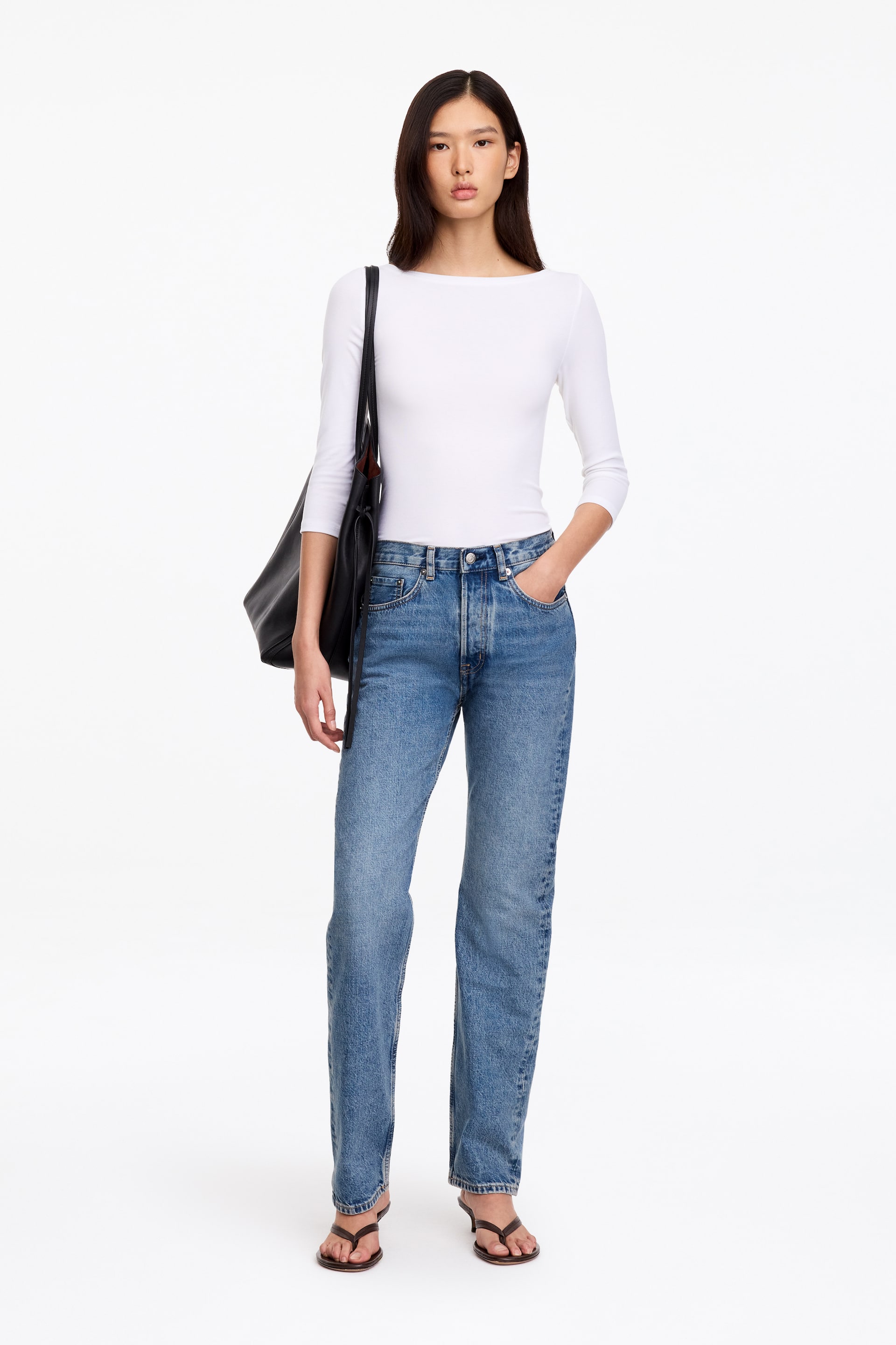 Grösseres Bild ansehen: Diese blaue Denim-Jeans mit mittlerer Bundhöhe und geradem Bein wird vom Model getragen, das mit einer Hand in der vorderen Tasche steht. Die Jeans ist mit einem figurbetonten weissen Langarm-Top und einer schwarzen Schultertasche kombiniert.
