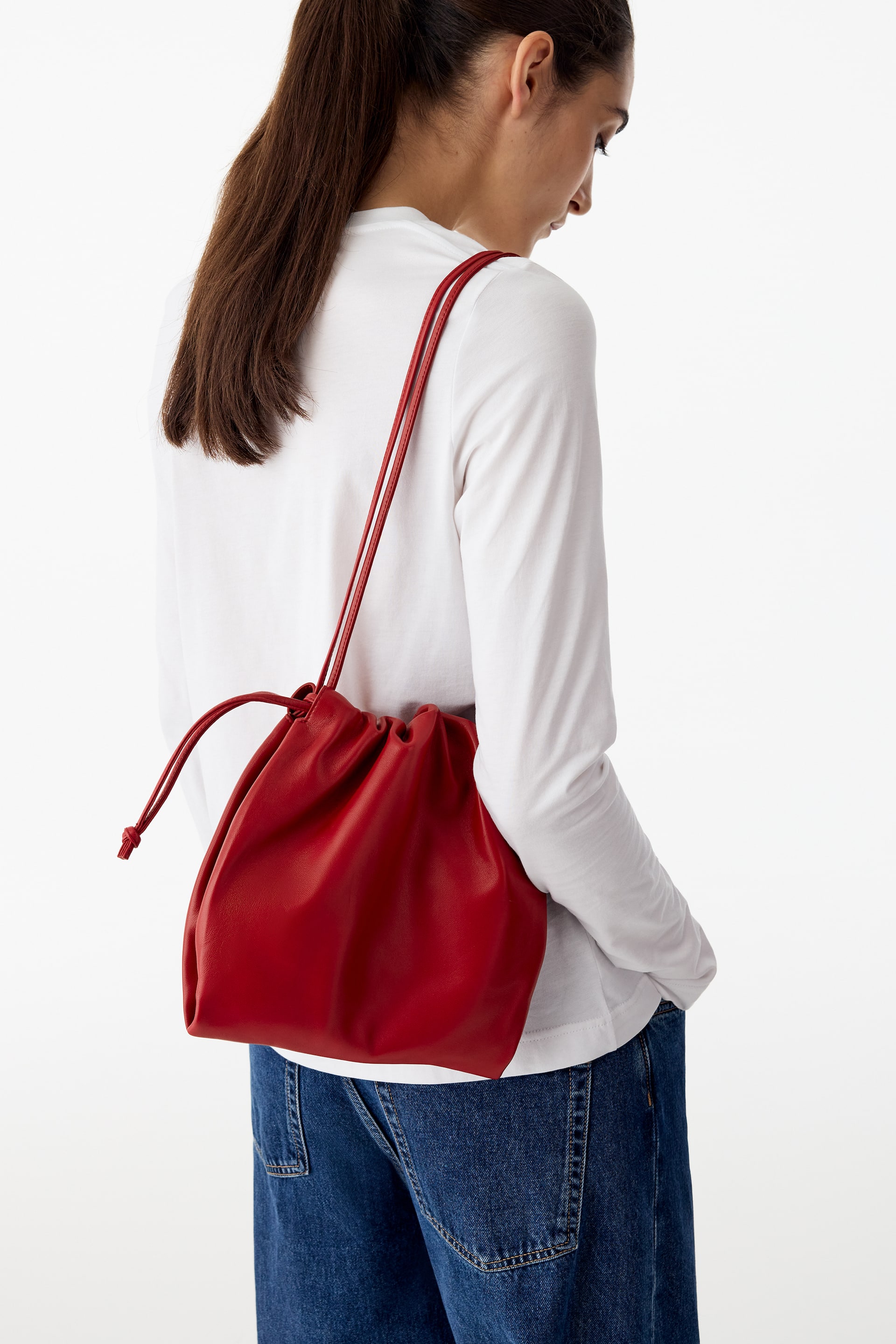 Agrandir l'image: Un sac en cuir rouge doté d’une ouverture froncée à cordon de serrage est porté à l’épaule par le modèle. Les fines bandoulières en cuir du sac reposent sur l’épaule du modèle, qui porte un haut blanc à manches longues et un jean bleu.