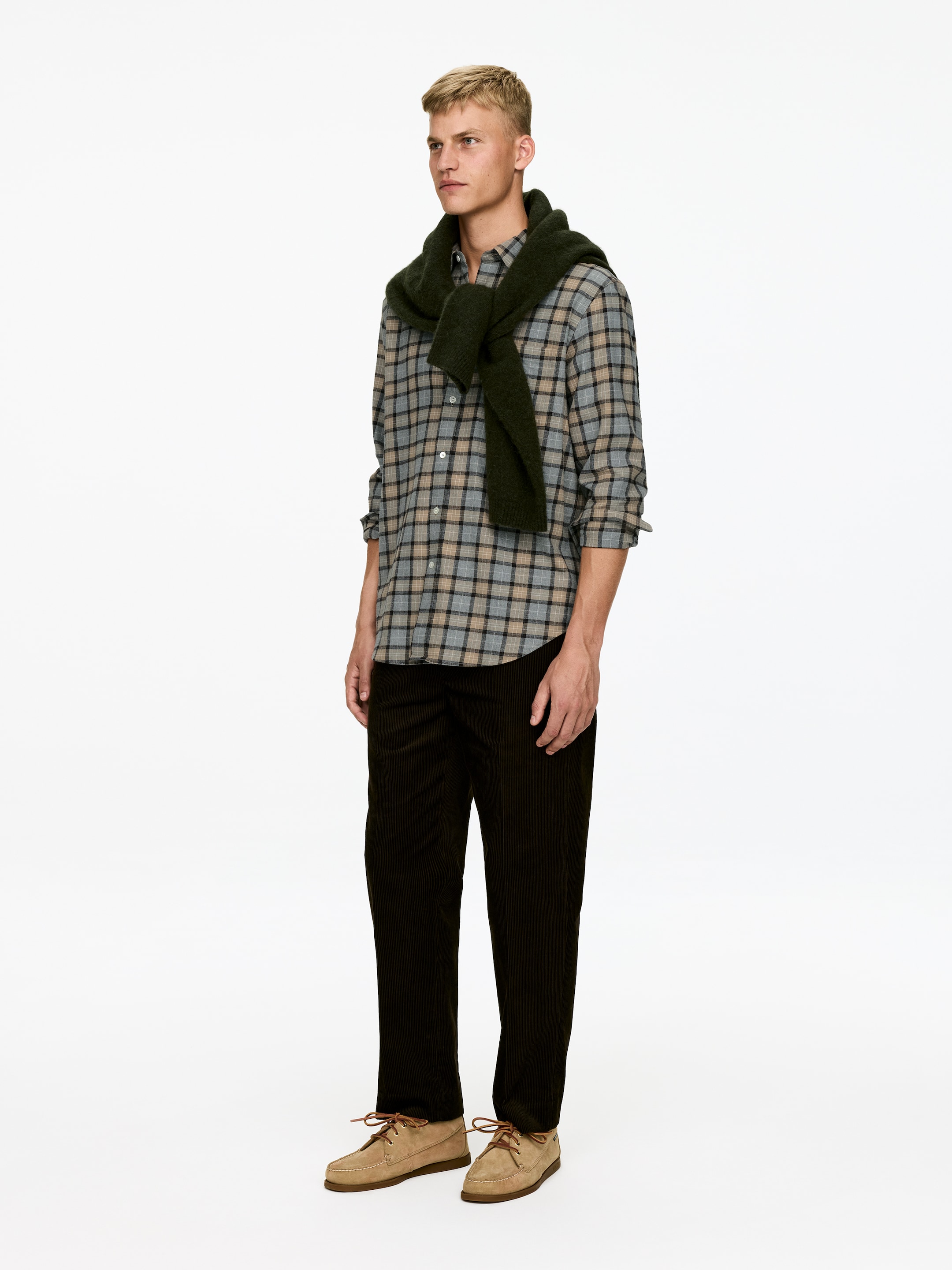 Ver imagen más grande: Un conjunto se presenta desde un ángulo de tres cuartos, con una camisa de botones de cuadros en gris, beige y negro, con un suéter verde oscuro colgado sobre los hombros. Unos pantalones de pana marrón oscuro y zapatos beige con cordones completan el conjunto.
