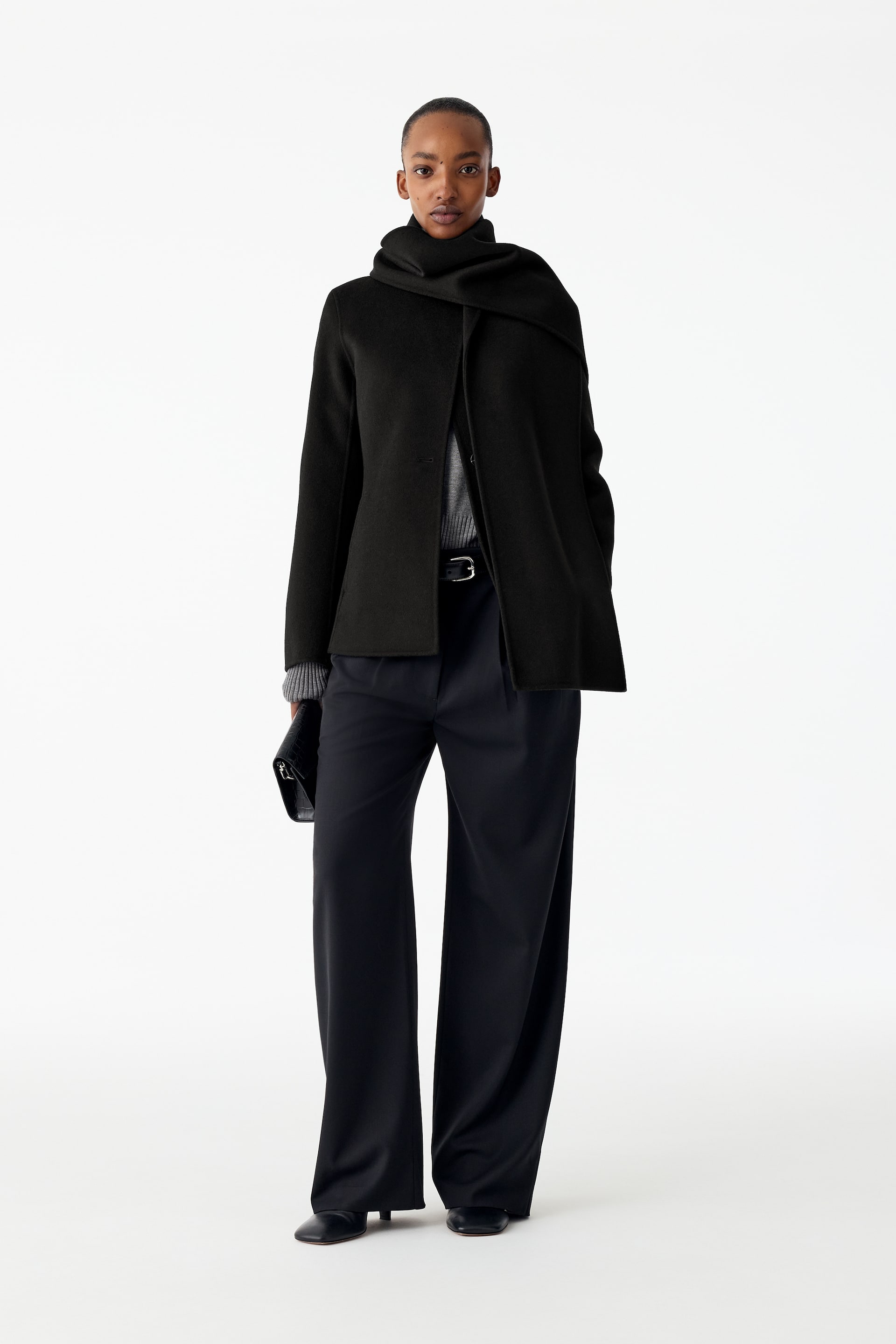 Agrandir l'image: Un blazer noir en mélange de laine, doté d’une écharpe amovible assortie enroulée autour du cou, est porté par le mannequin. Le blazer cintré est superposé à un pull en maille gris et à un pantalon foncé, tandis qu’une pochette noire est tenue dans une main.