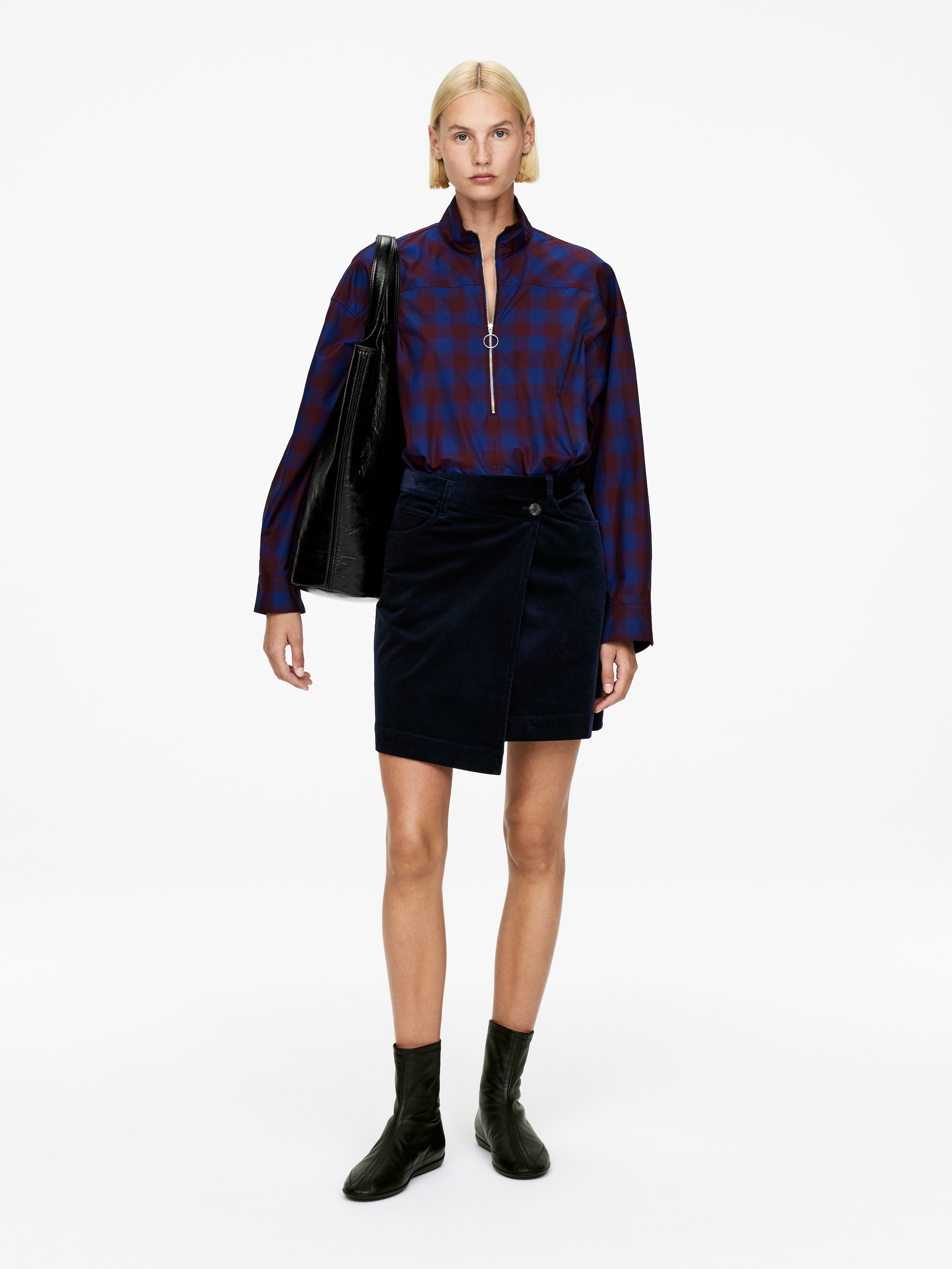 Visualizza immagine più grande: Una camicia a maniche lunghe, caratterizzata da un motivo a quadri rosso e blu navy, un colletto alla coreana e una chiusura a mezza zip con un tirazip circolare, è indossata infilata in una minigonna di velluto a coste blu navy. La gonna è progettata con un davanti stile avvolgente e chiusura a bottoni. Stivaletti neri alla caviglia con finitura liscia e gambale aderente sono anch'essi indossati, e una borsa tote nera è portata a spalla.