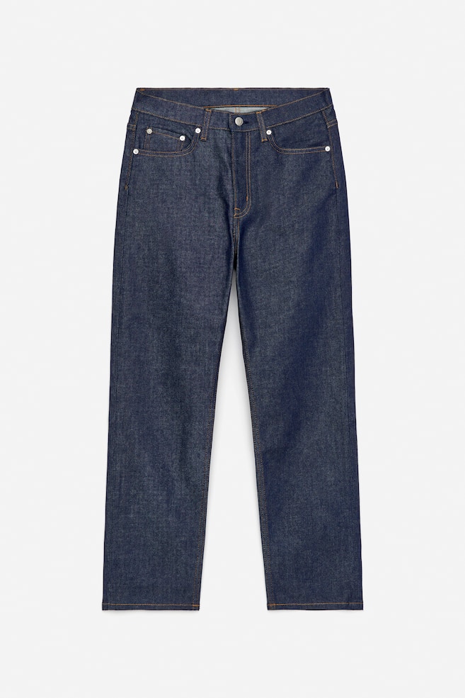 Ein Paar dunkel indigofarbene Jeans wird von vorne präsentiert, mit einer mittelhohen Taille, klassischem Fünf-Taschen-Design und einem Reißverschluss mit Knopfverschluss. Kontrastierende orangefarbene Nähte und silberfarbene Nieten sind sichtbar, wobei die Beine schlank, konisch und gekürzt erscheinen. - 1