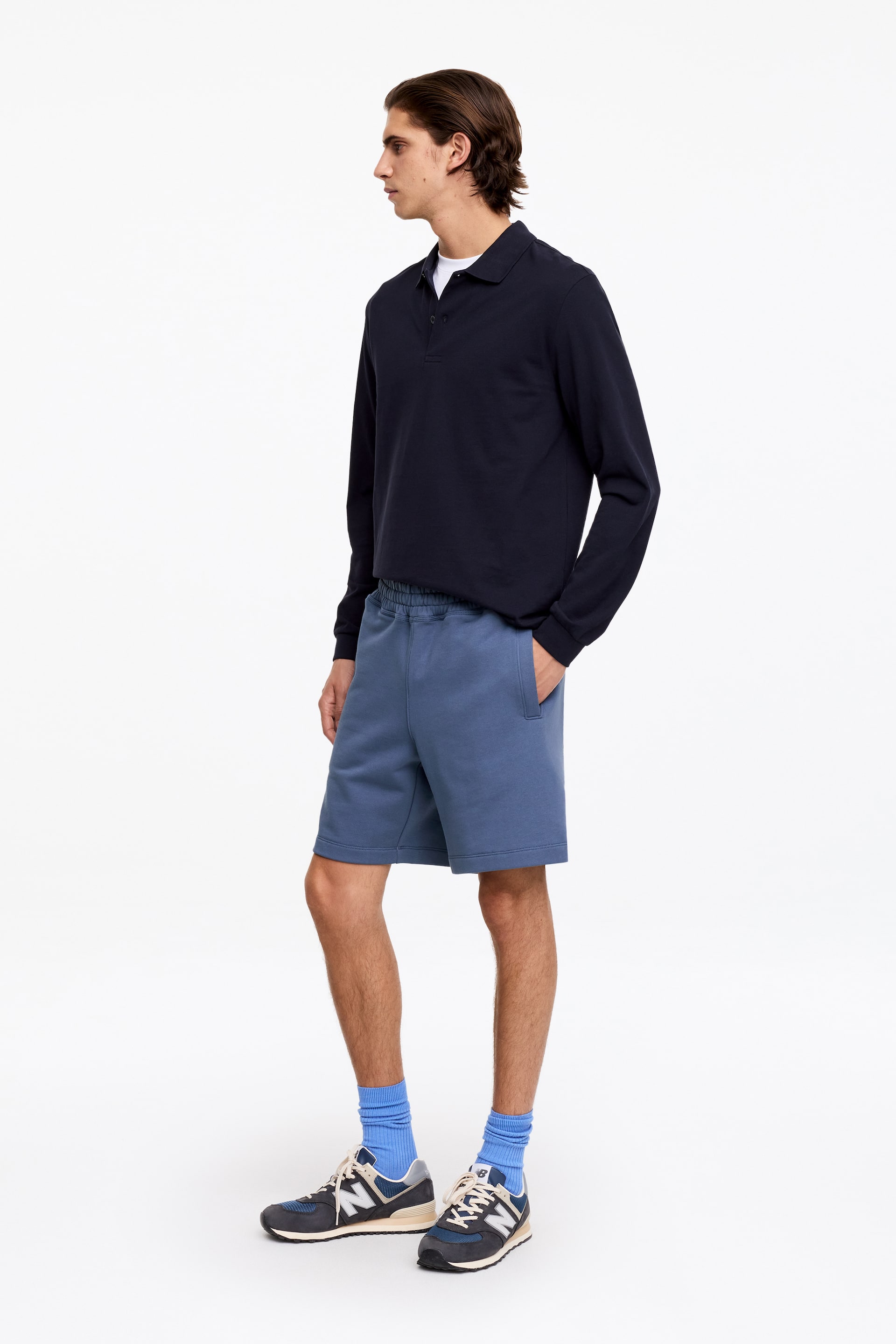 Agrandir l'image: Le mannequin porte des shorts bleus sans effort, confectionnés en molleton français. Ils sont dotés d'une taille élastiquée et de poches passepoilées visibles sur les coutures latérales, et la main du mannequin repose dans l'une des poches latérales.