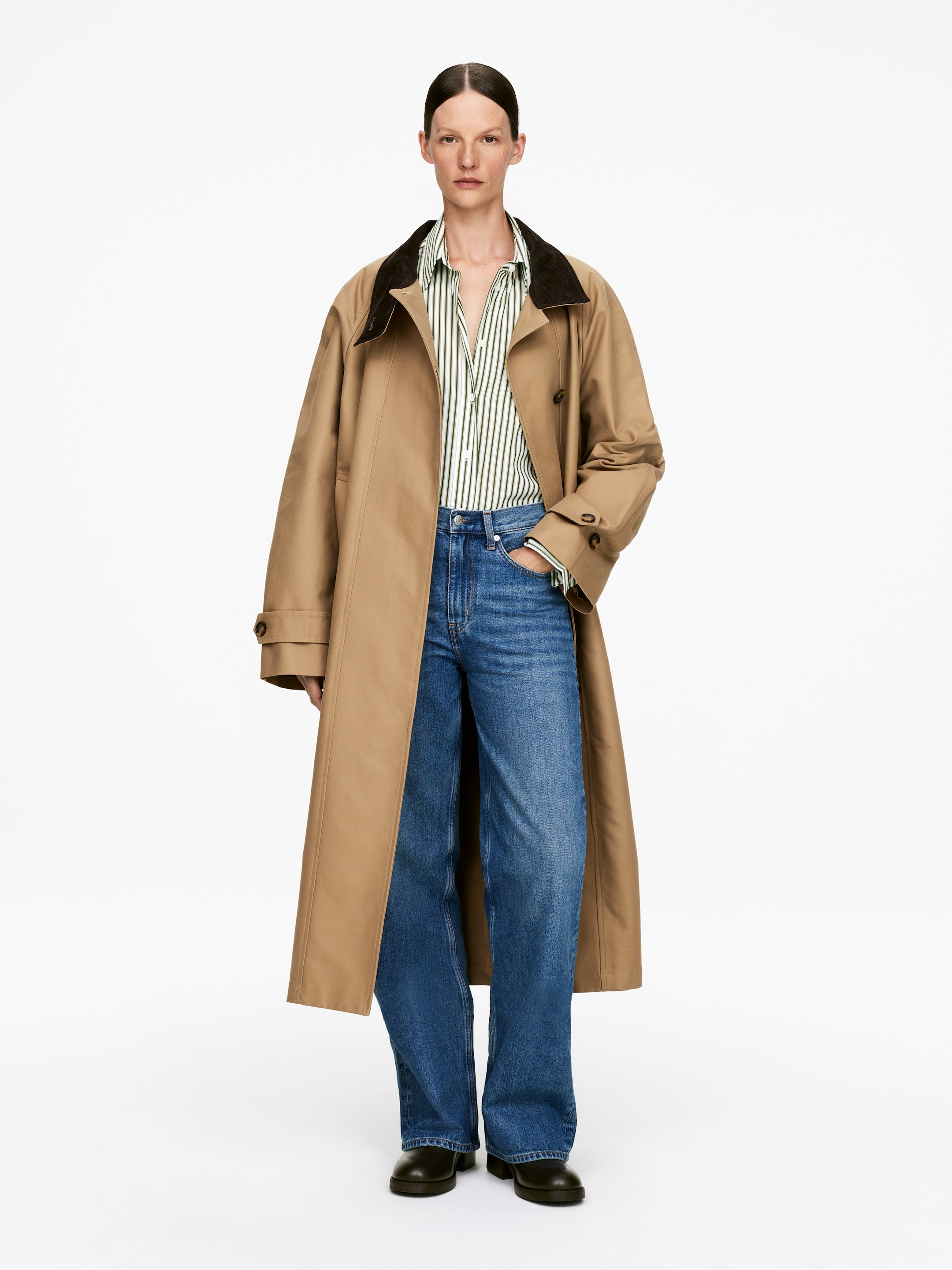 Agrandir l'image: Un trench-coat croisé de couleur fauve est porté ouvert, avec un col contrastant brun foncé, des manches larges avec des poignets ajustables à boutons, et des boutons visibles à l'avant. Il est superposé à une chemise à rayures blanches et vertes, un jean bleu à jambes larges, et des chaussures sombres à semelles épaisses.
