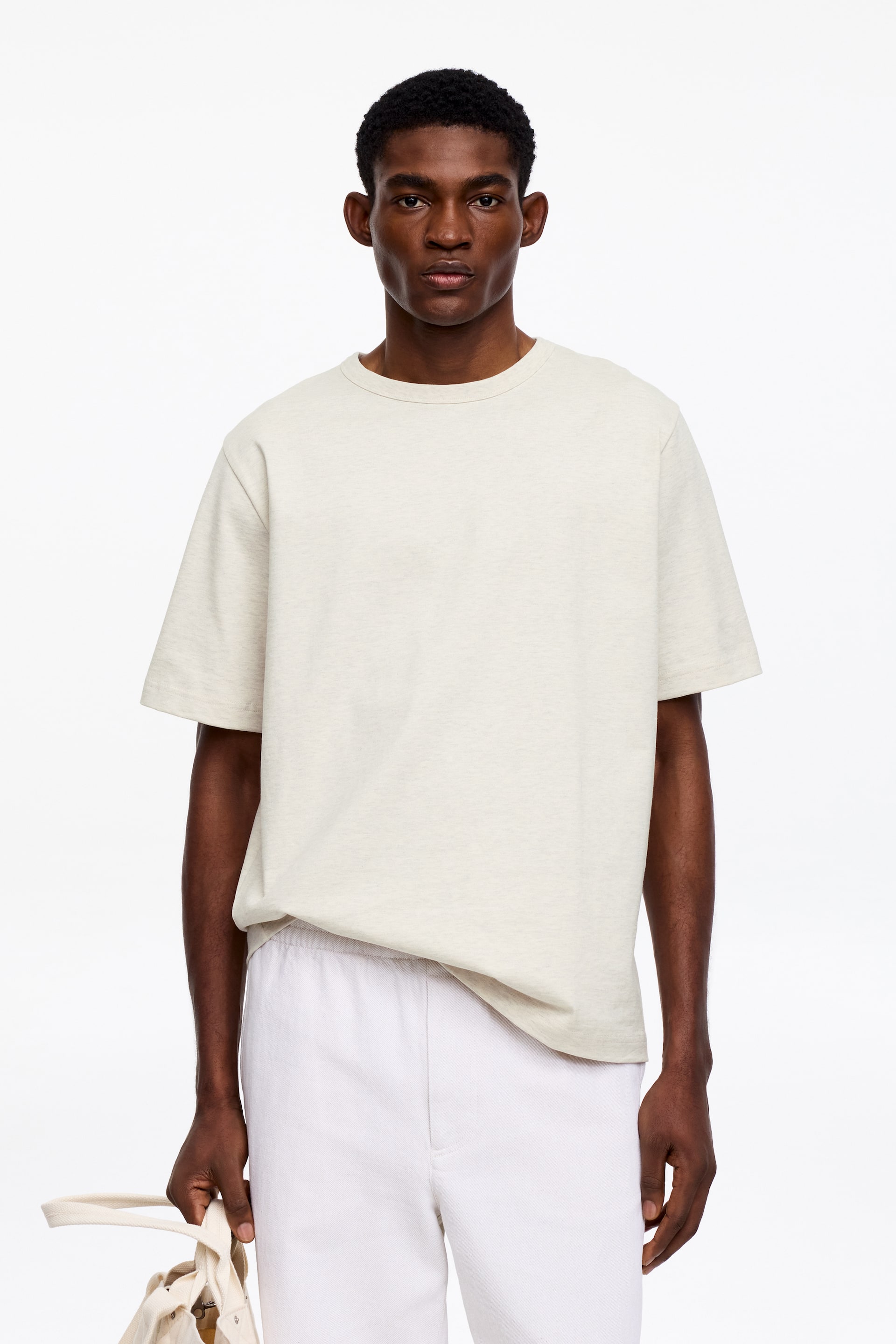 Agrandir l'image: Un t-shirt beige clair chiné présente une coupe ample et décontractée à l'avant, mettant en valeur un col rond côtelé et des manches courtes, avec son ourlet partiellement rentré dans un pantalon blanc.