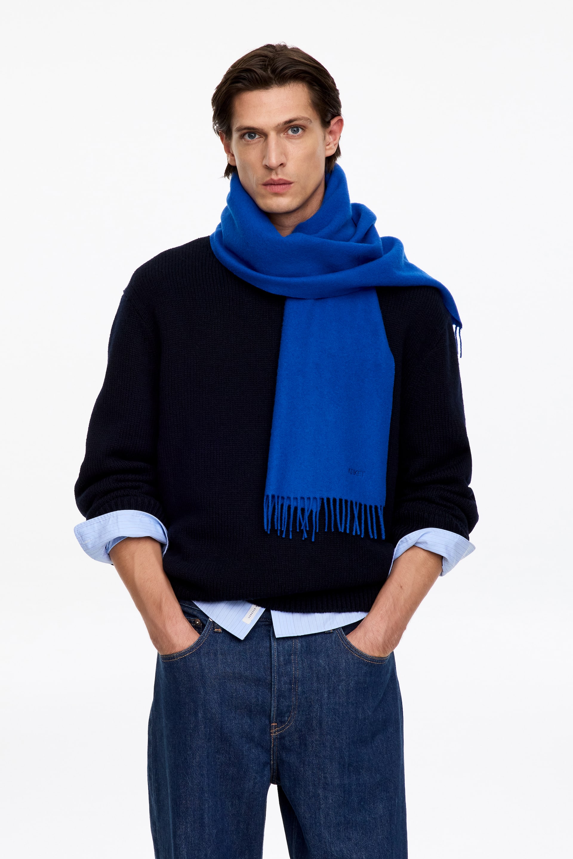 View larger image: A bright blue scarf with fringed ends is worn wrapped around the model’s neck, with one end hanging down the front. The scarf is layered over a dark navy-blue knit sweater, a light blue collared shirt, and dark blue jeans.