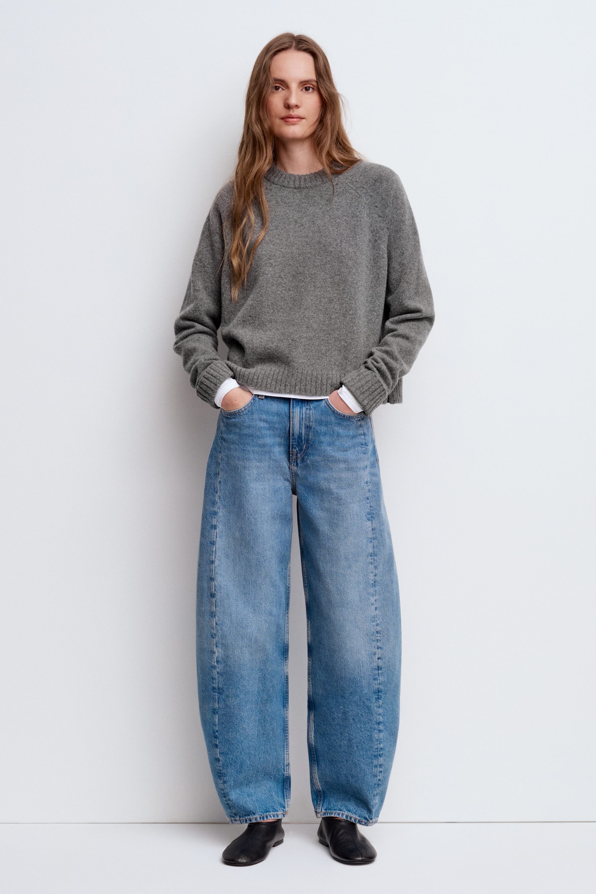 Visa större bild: Framsidan av en grå sweater med rund hals, med avslappad passform, långa ärmar med ribbade muddar och ribbad nederkant, bärs lager på lager över en vit skjorta. Den stylas med blå jeans med vida ben som har en tydlig vertikal söm på varje ben, samt svarta platta slip-on-skor.