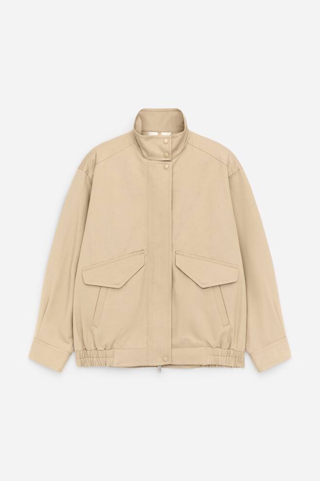Een beige Harringtonjas wordt van voren gepresenteerd, met een opstaande kraag met drukknopen, duidelijke stiksels op de schouders en een voorflap die een ritssluiting verbergt, zichtbaar aan een ritstrekker onderaan. De jas is uitgevoerd met twee grote opgestikte zakken met knoopsluitende kleppen, manchetten met knopen en elastische details aan de zijzomen. - 1