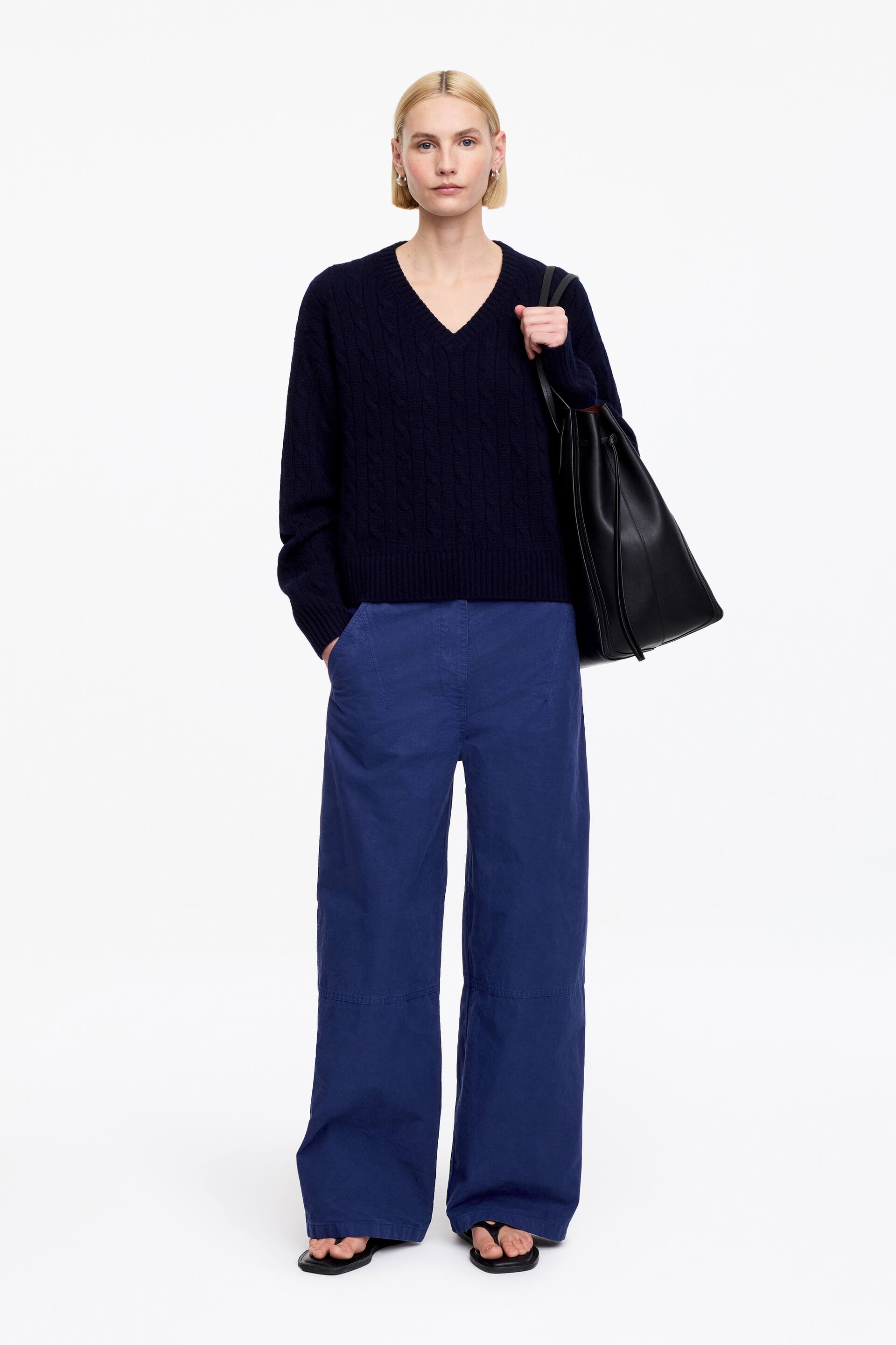 Ver imagen más grande: Se presenta un conjunto completo, de frente, que incluye un jersey de punto trenzado azul marino con cuello en V y puños y dobladillo acanalados, pantalones anchos azul real con detalles de costura en las rodillas y bolsillos laterales, y sandalias de dedo negras, llevando un bolso tote negro suave en el hombro izquierdo.