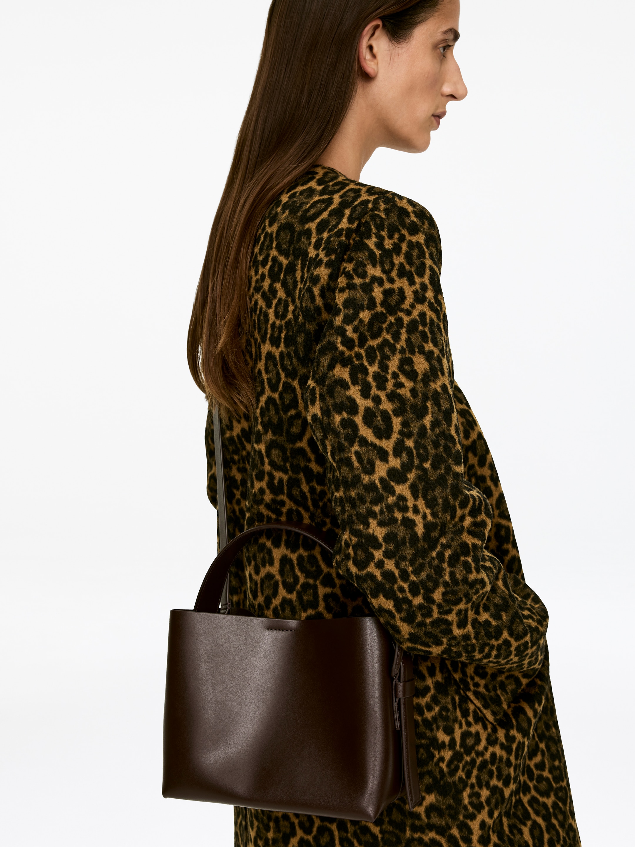View larger image: A mid-size, rigid dark brown crossbody bag is being carried on the model's shoulder. The bag features a wide top handle, and its adjustable strap is visible over a leopard print garment.