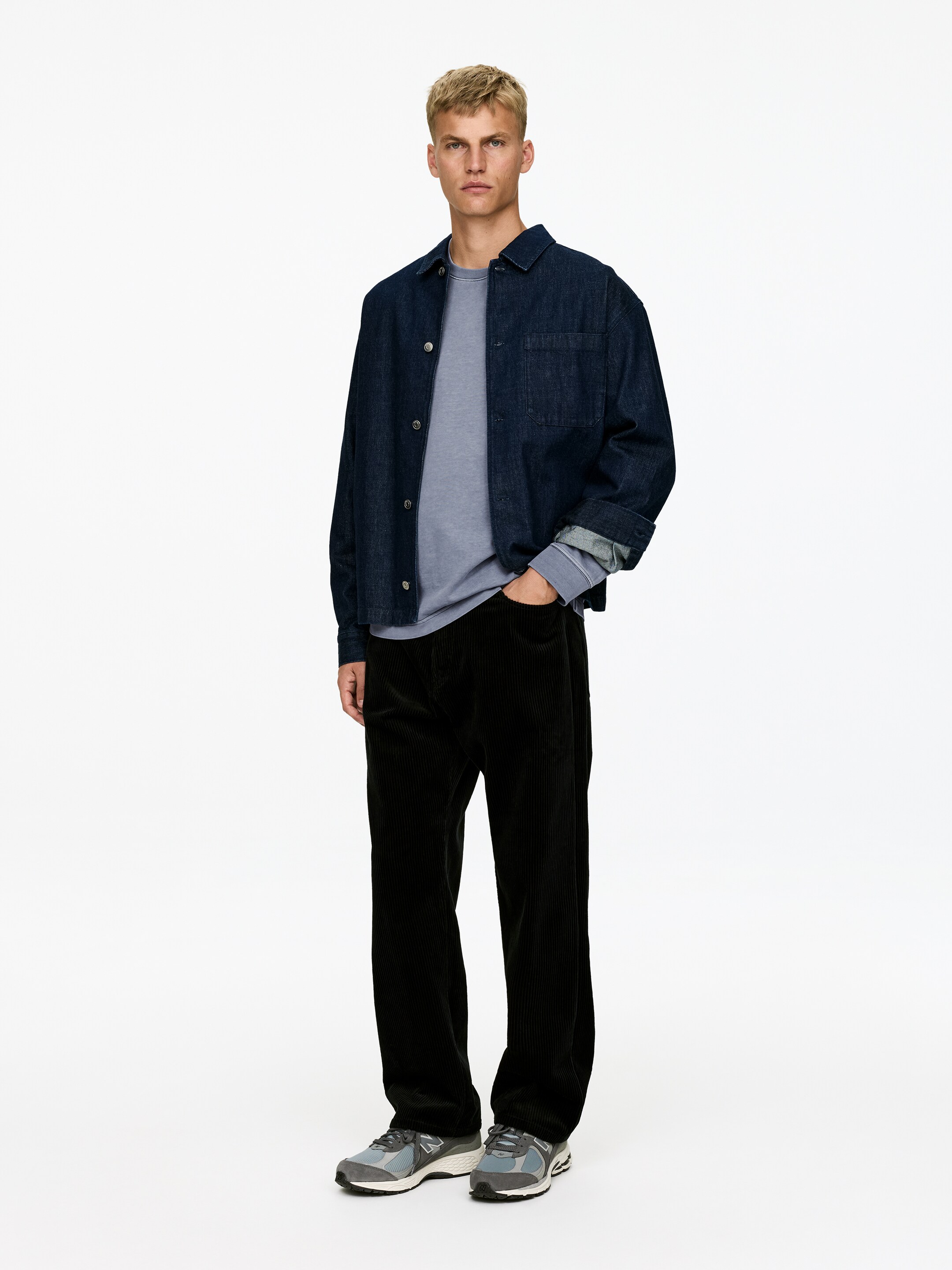 View larger image: An outfit is being presented from a front-facing angle, featuring a dark blue denim overshirt with rolled-up sleeves and a visible chest pocket, worn over a light blue long-sleeved crewneck shirt, with black corduroy wide-leg pants and grey and blue sneakers completing the ensemble.