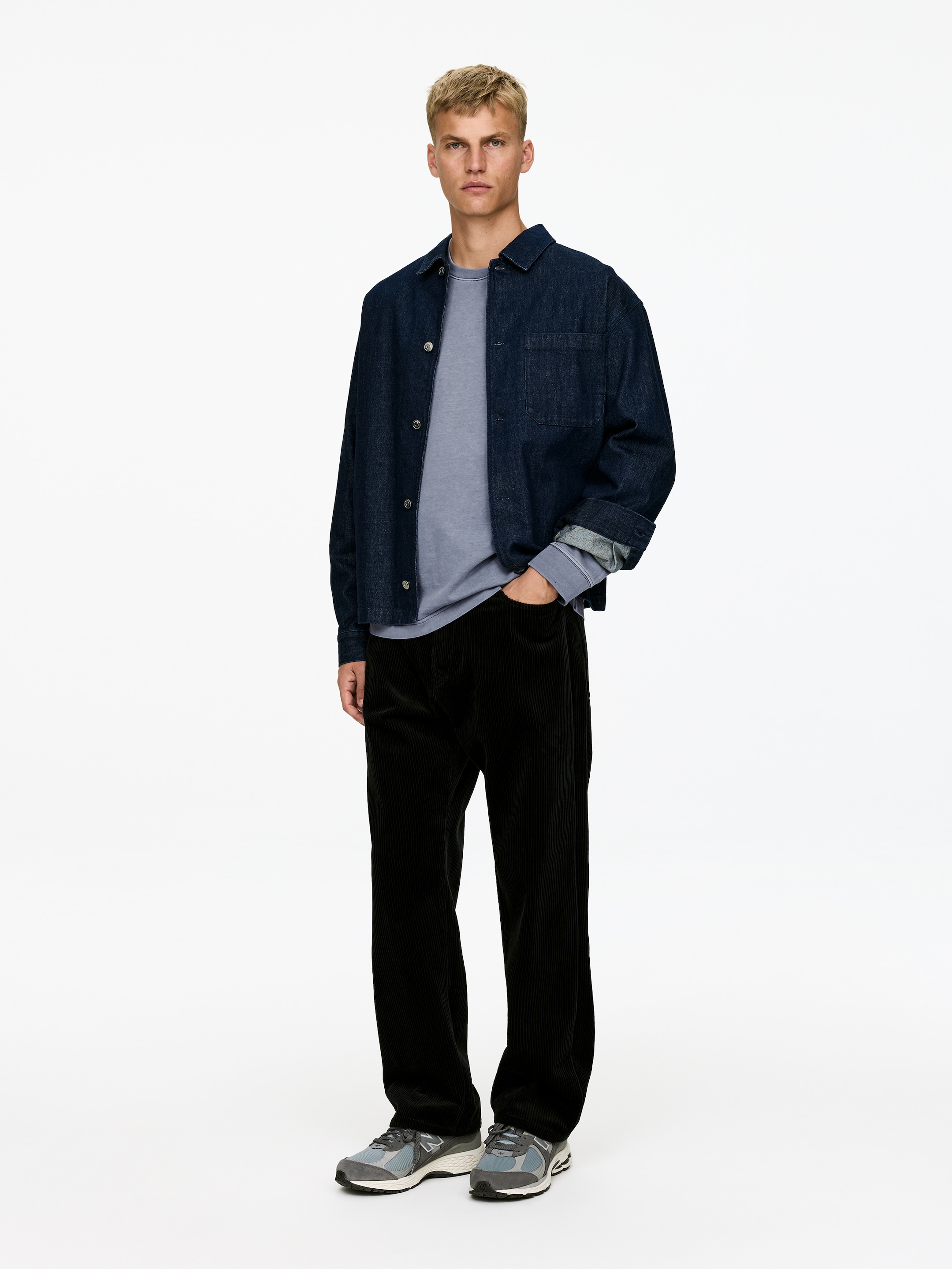 View larger image: An outfit is being presented from a front-facing angle, featuring a dark blue denim overshirt with rolled-up sleeves and a visible chest pocket, worn over a light blue long-sleeved crewneck shirt, with black corduroy wide-leg pants and grey and blue sneakers completing the ensemble.