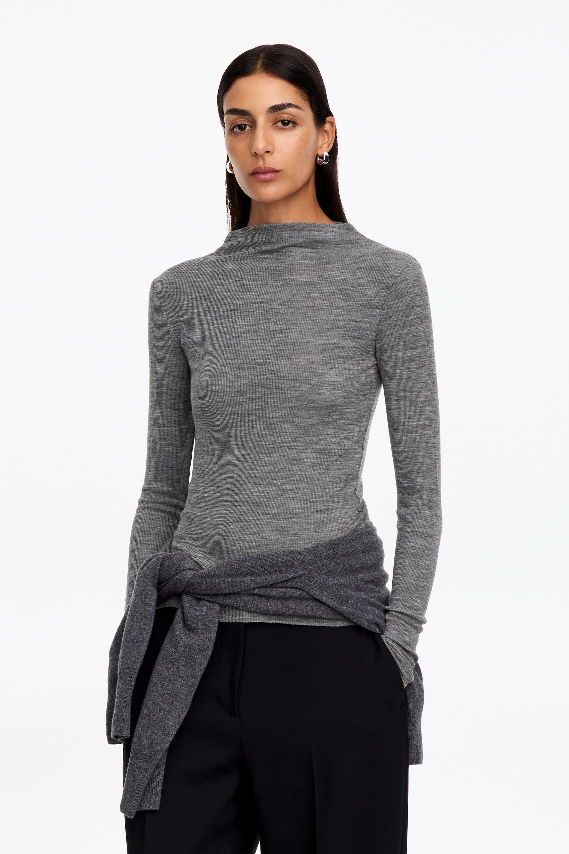 Vezi imaginea mai mare: Un top gri melanj, din tricot fin, cu mânecă lungă și guler înalt tip mock neck, este purtat de model. Un pulover din tricot gri închis este legat în jurul taliei peste top, care este asortat cu pantaloni negri.